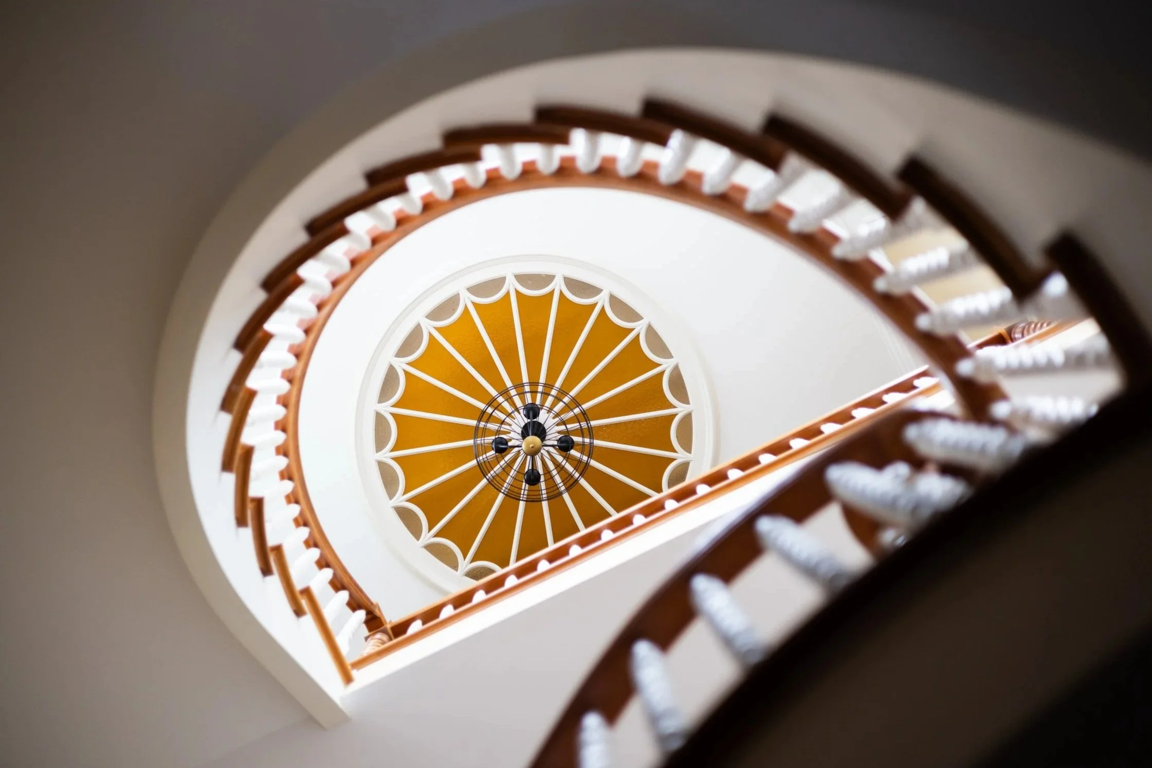 Looking up a spiral staircase with wooden handrails and white balusters towards a ceiling with a decorative circular pattern and a central chandelier.