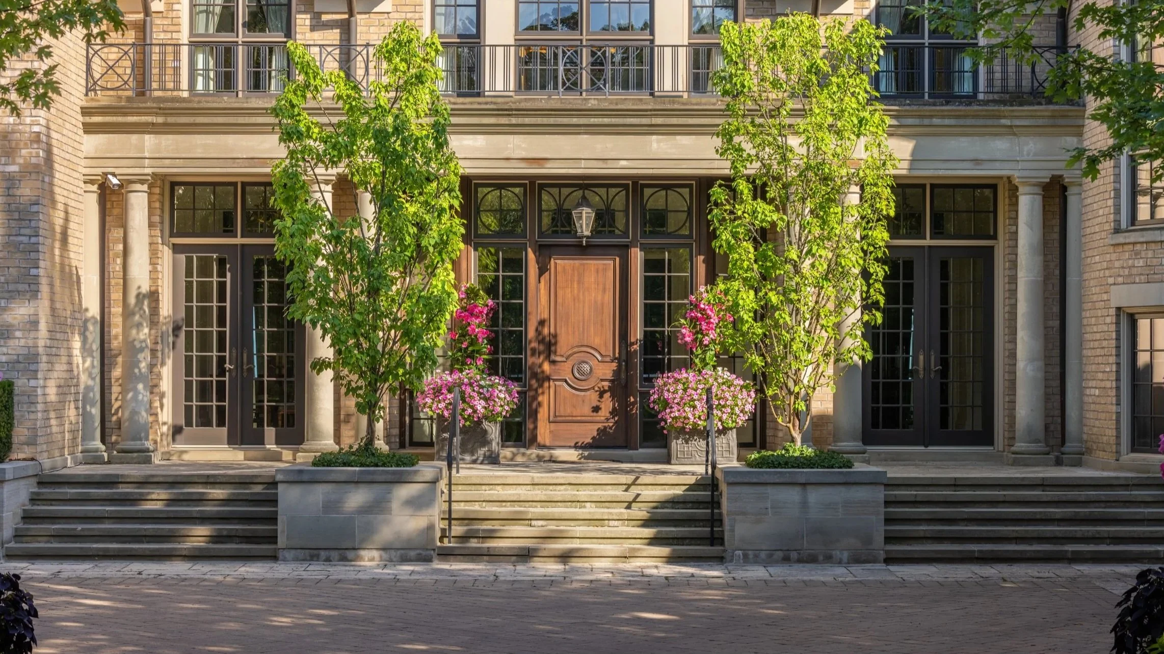 $40m home. Front entrance of a building with steps, two trees, pink flowers in pots, and large glass doors.