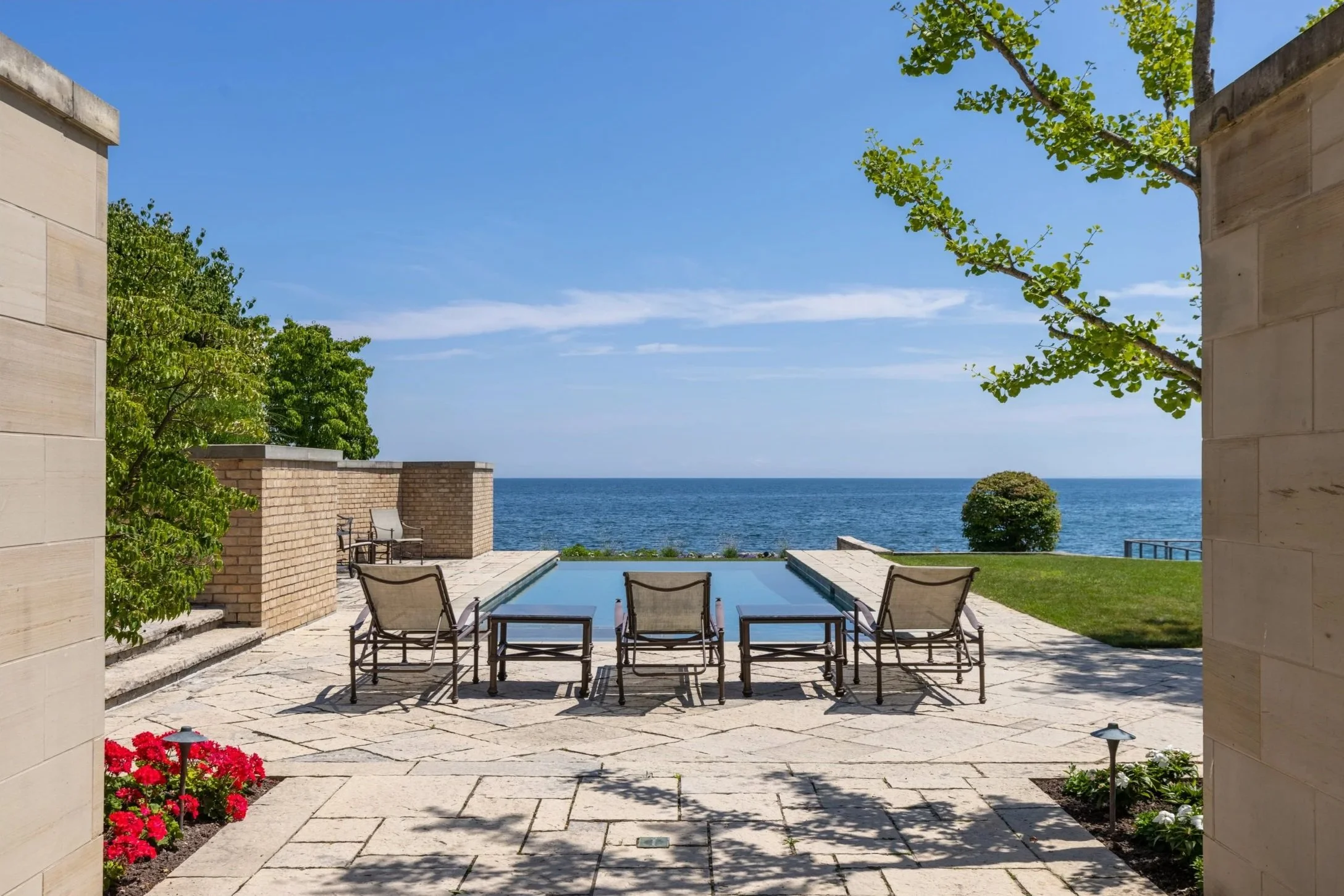 View of a backyard patio with a small pool, lawn, and ocean beyond, with four chairs around the pool and trees and flowers nearby.