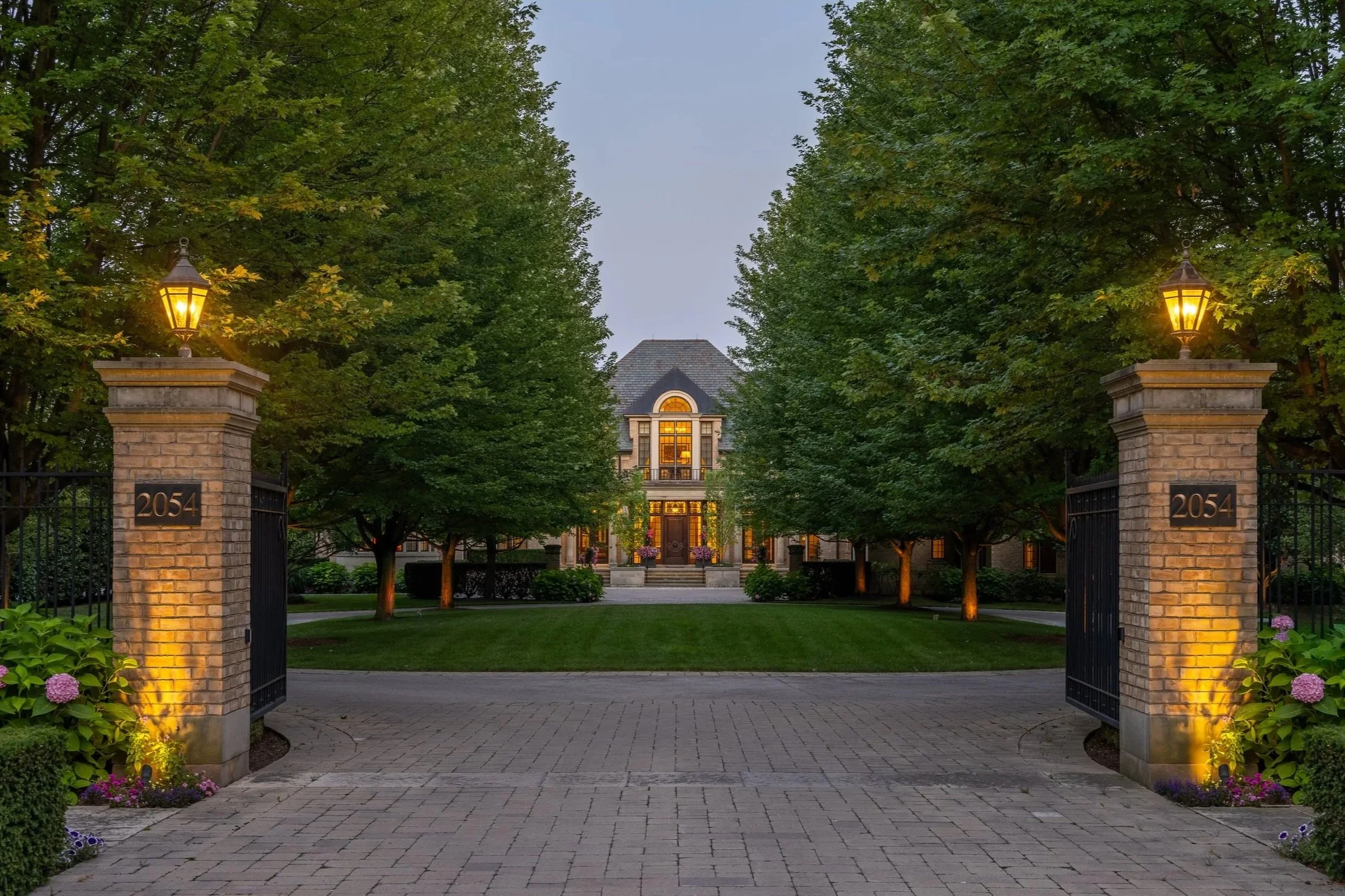 A large house with a gray roof and large windows, surrounded by green trees and a grassy lawn, viewed from the driveway at dusk, with two lit lanterns on brick pillars at the gate.