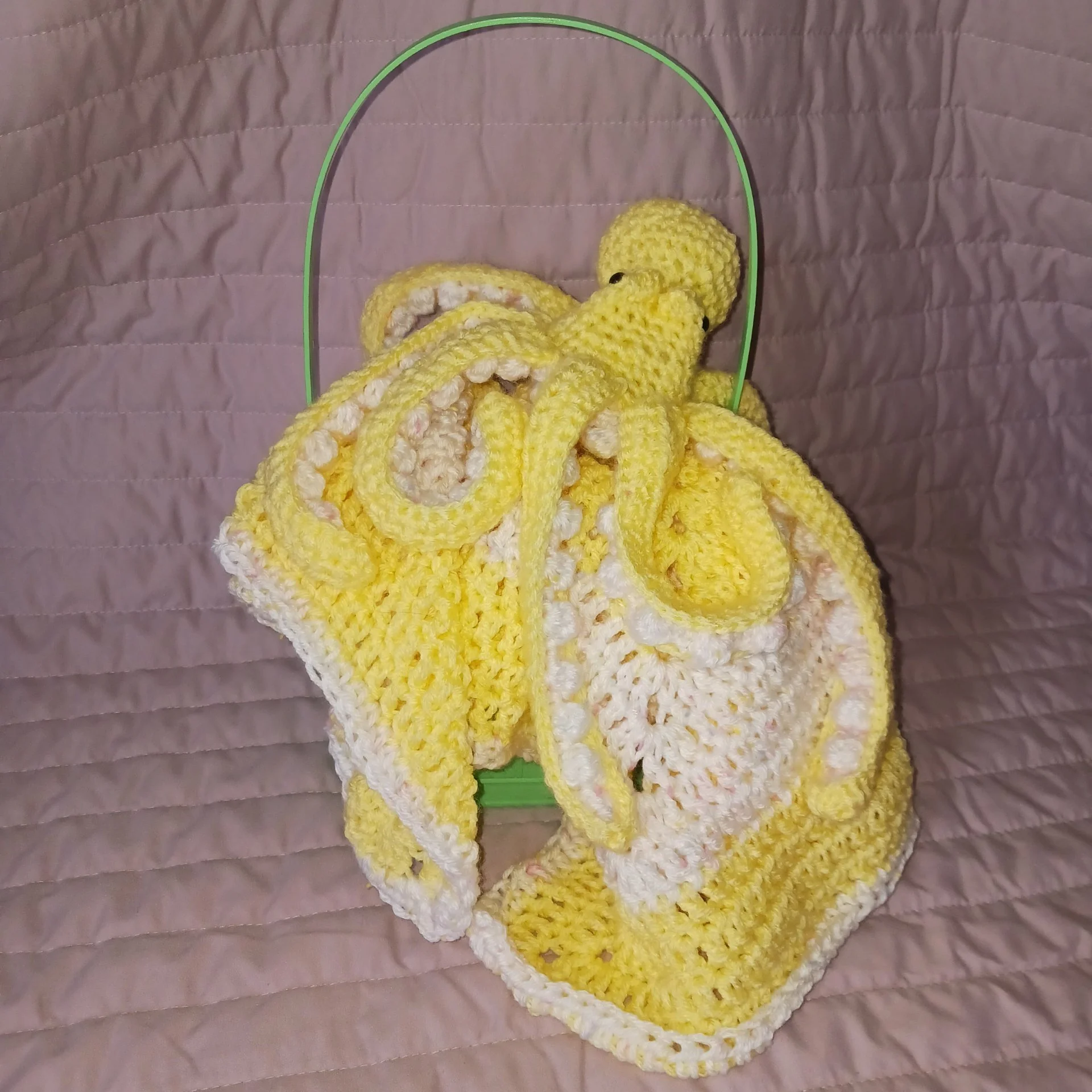 A crocheted yellow octopus with black eyes, surrounded by crocheted yellow and white tentacles, hanging inside a green and metal wire frame, all placed on a quilted pink surface.