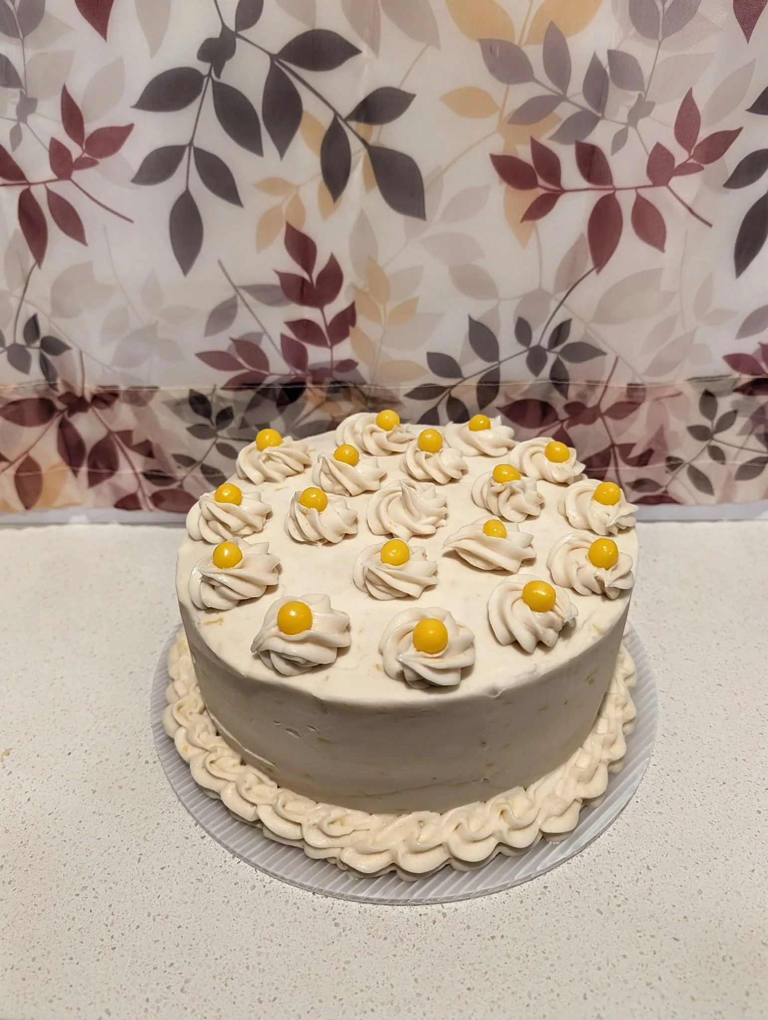 A round white cake decorated with white frosting swirls and yellow candies, sitting on a white surface with a wallpapered background featuring a leaf pattern.