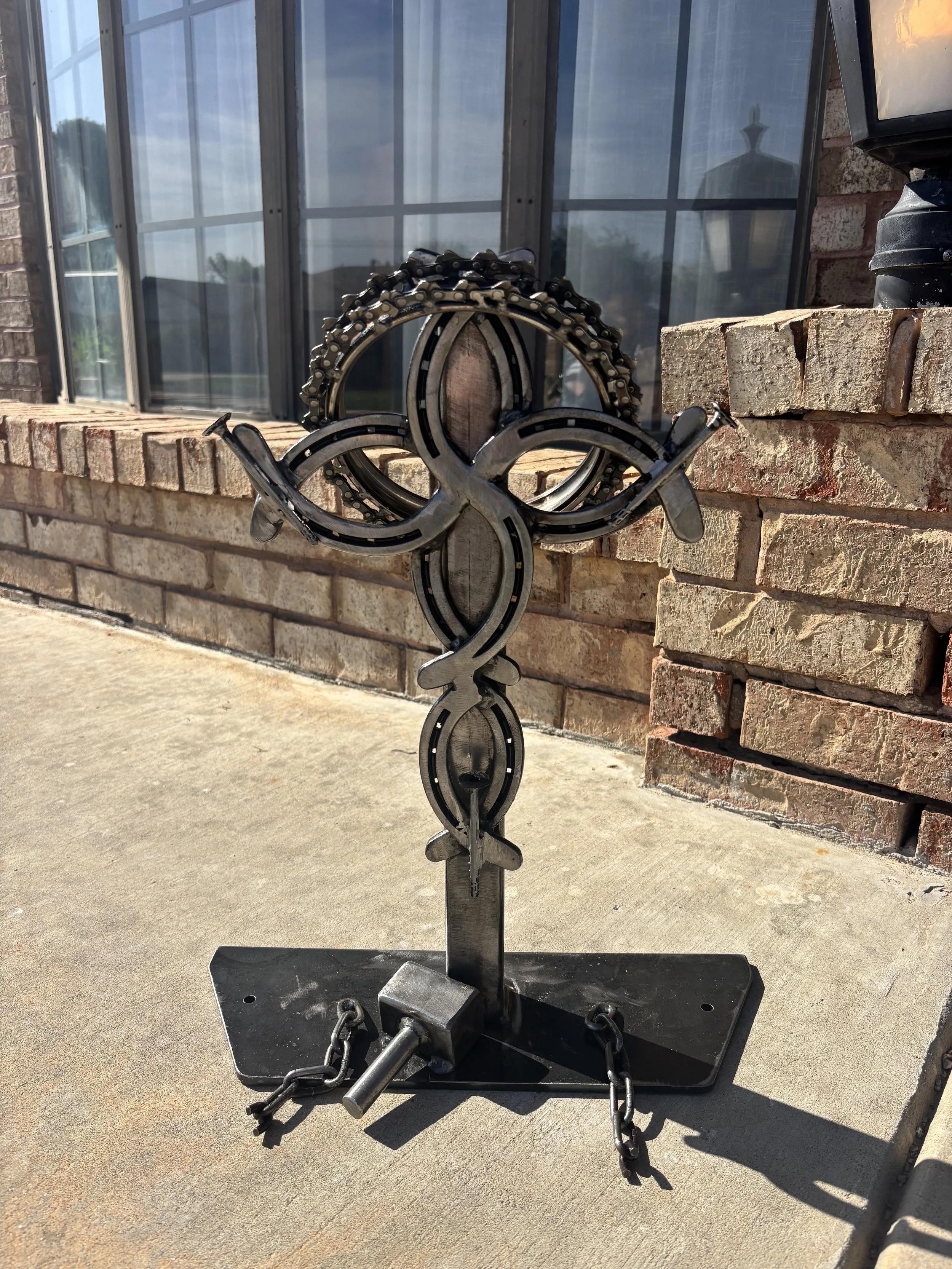 A metal sculpture of a fish with a tall fin, made from bike chains, mounted on a stand outside on a concrete sidewalk with a brick wall and window in the background.