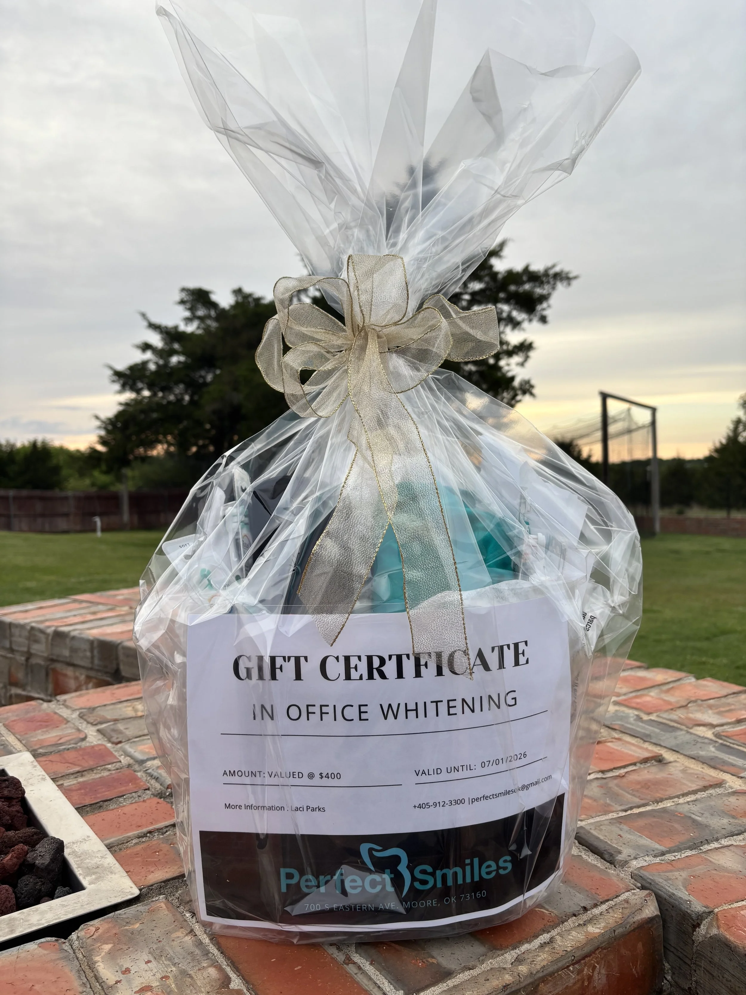 Gift certificate in clear plastic wrapping with a gold bow, placed on a brick surface outdoors with trees and a hockey goal in the background.