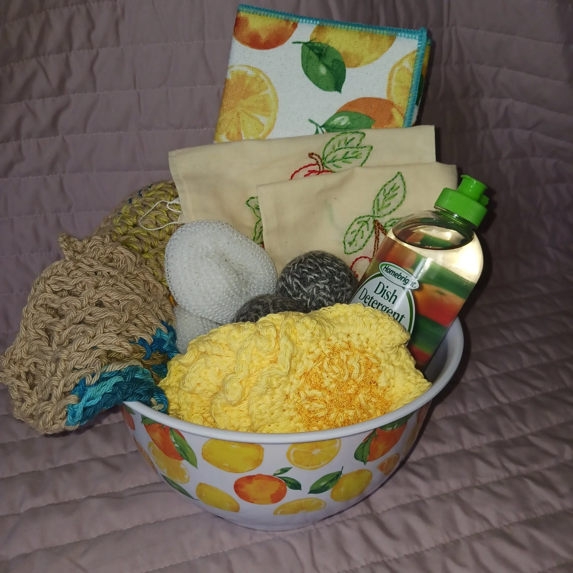 A white laundry basket with lemon and orange fruit pattern, filled with knitted and embroidered washcloths, crochet and knitted items, and a bottle of dish detergent.
