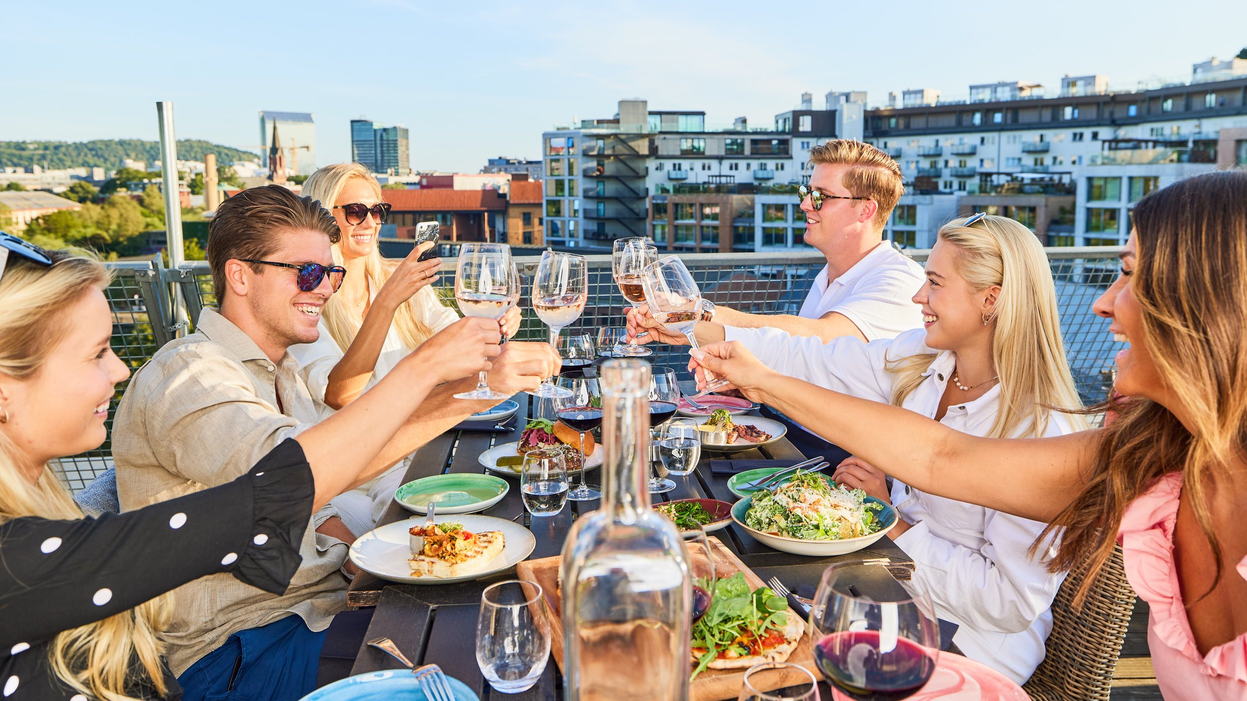 Four happy people smiling and toasting on the rooftop terrace of BAR Vulkan, seated at a table set with good food and drinks