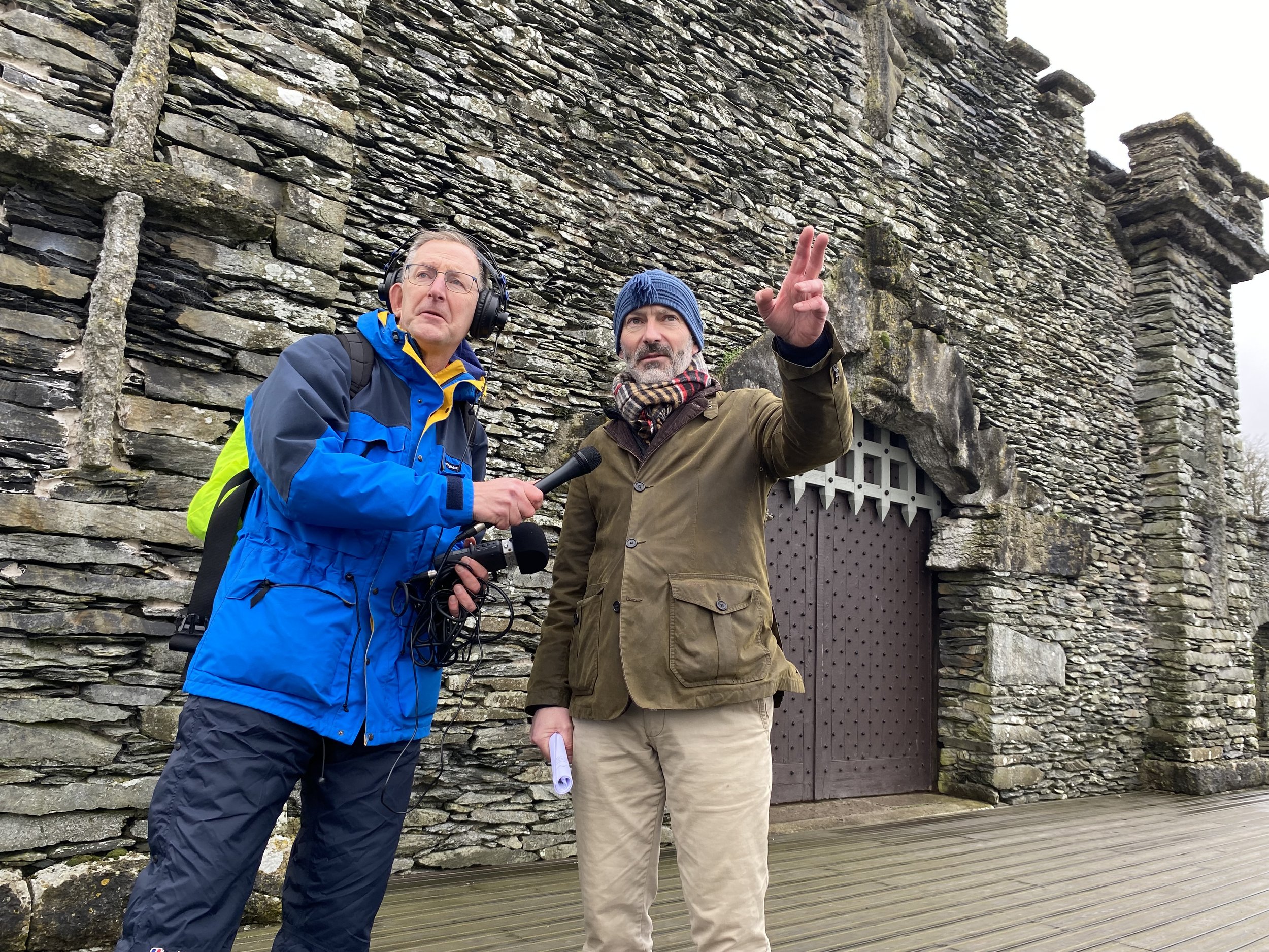 Two men outdoors in front of a stone wall, one holding a microphone, one pointing, dressed in jackets, one in a blue beanie and the other with glasses and headphones.