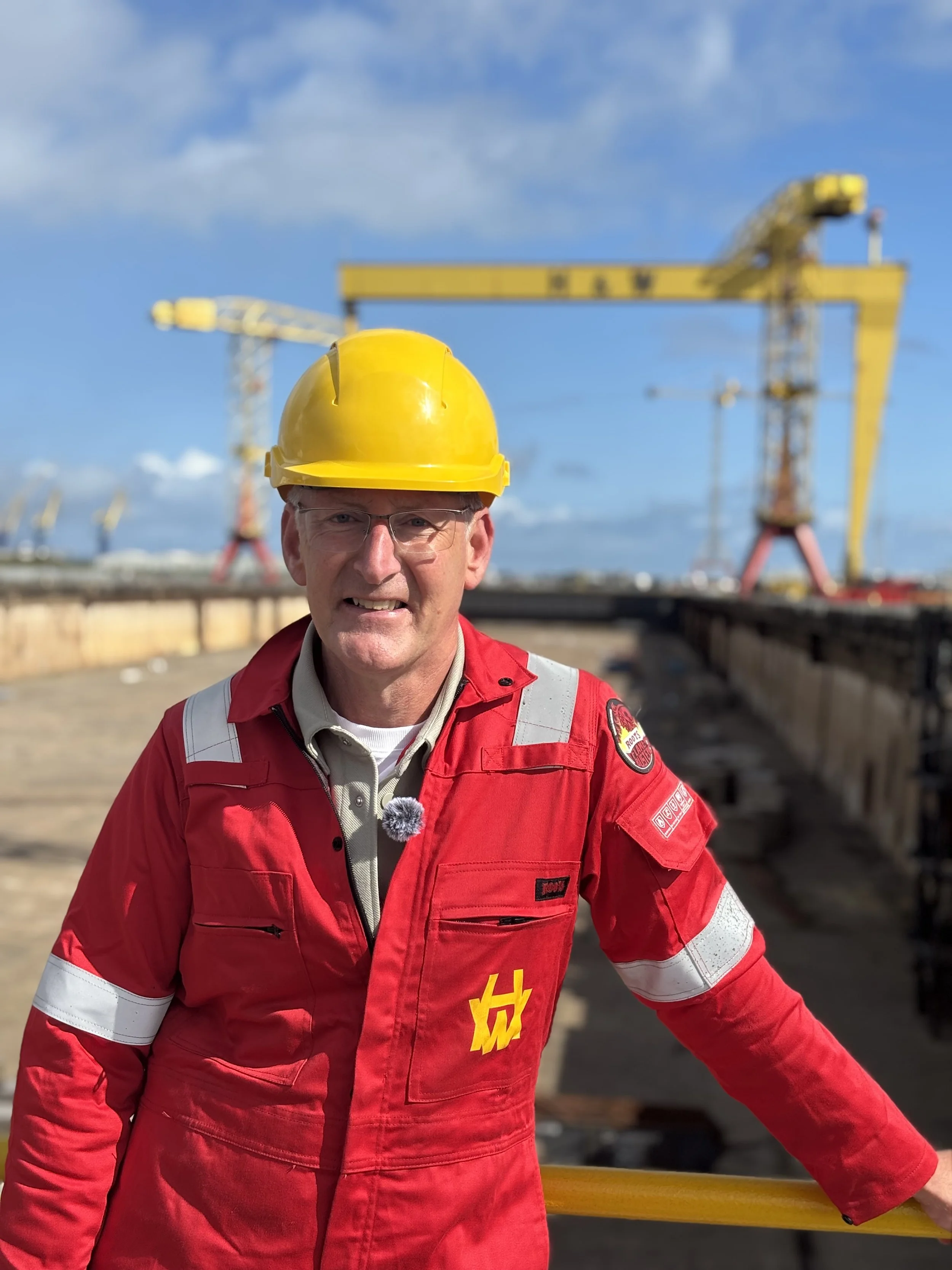 A man wearing a yellow safety helmet and a red safety jacket standing on a construction site with yellow cranes in the background.