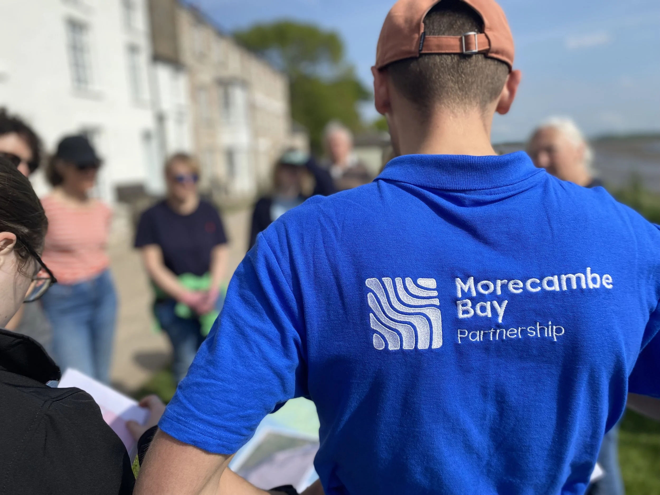 Close-up of a man wearing a blue shirt with the text 'Morecambe Bay Partnership' on the back, standing outdoors in a group of people with buildings and a tree in the background.