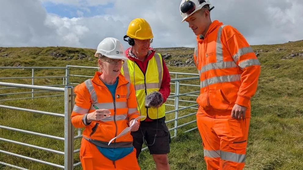 A woman in orange safety gear holds a headlamp and a paper, talking to a man in a red jacket and black shorts who is adjusting his headset outside a building with large glass windows.
