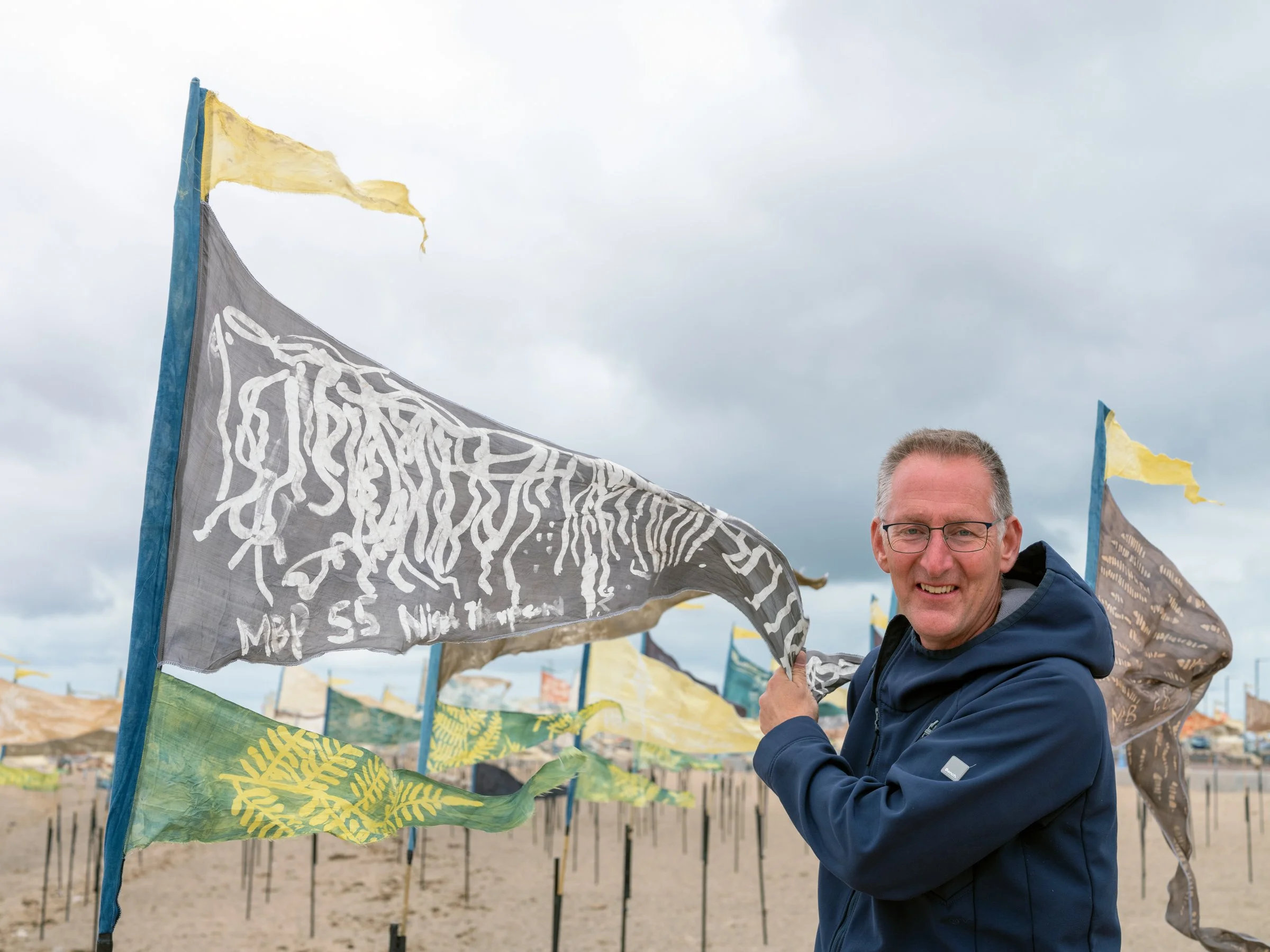 A man with glasses smiling at the camera, standing outdoors on a cloudy day, behind a row of colorful flags on poles.
