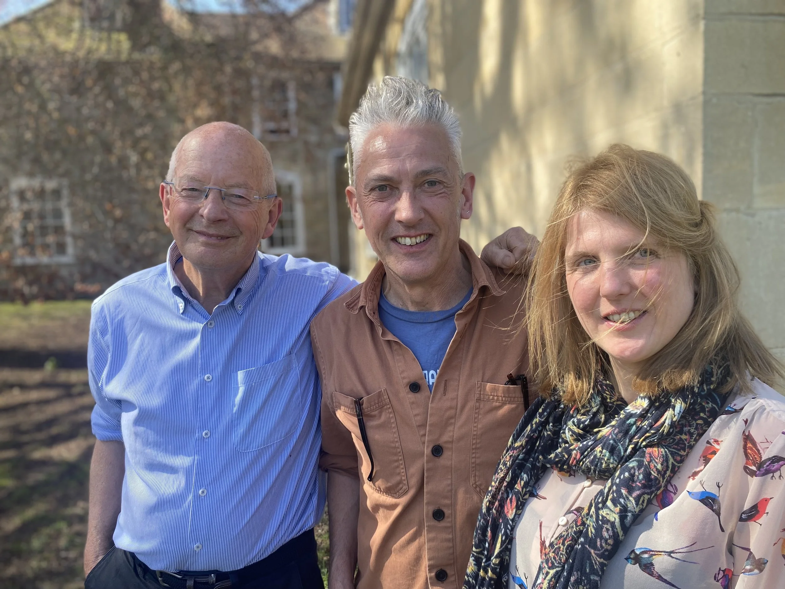 Four people smiling outdoors on a sunny day; two men and two women, standing close together, with trees and building in the background.