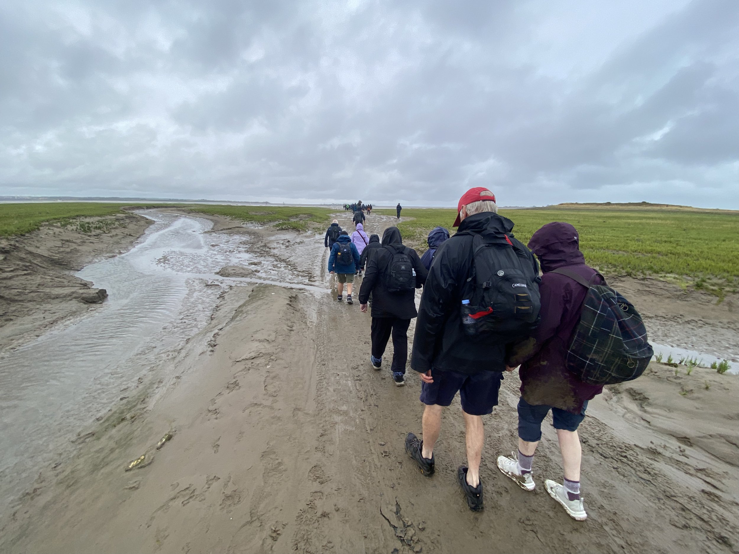 A group of people, dressed in outdoor clothing and backpacks, walking along a muddy, water-filled trail through a grassy landscape under a cloudy sky.
