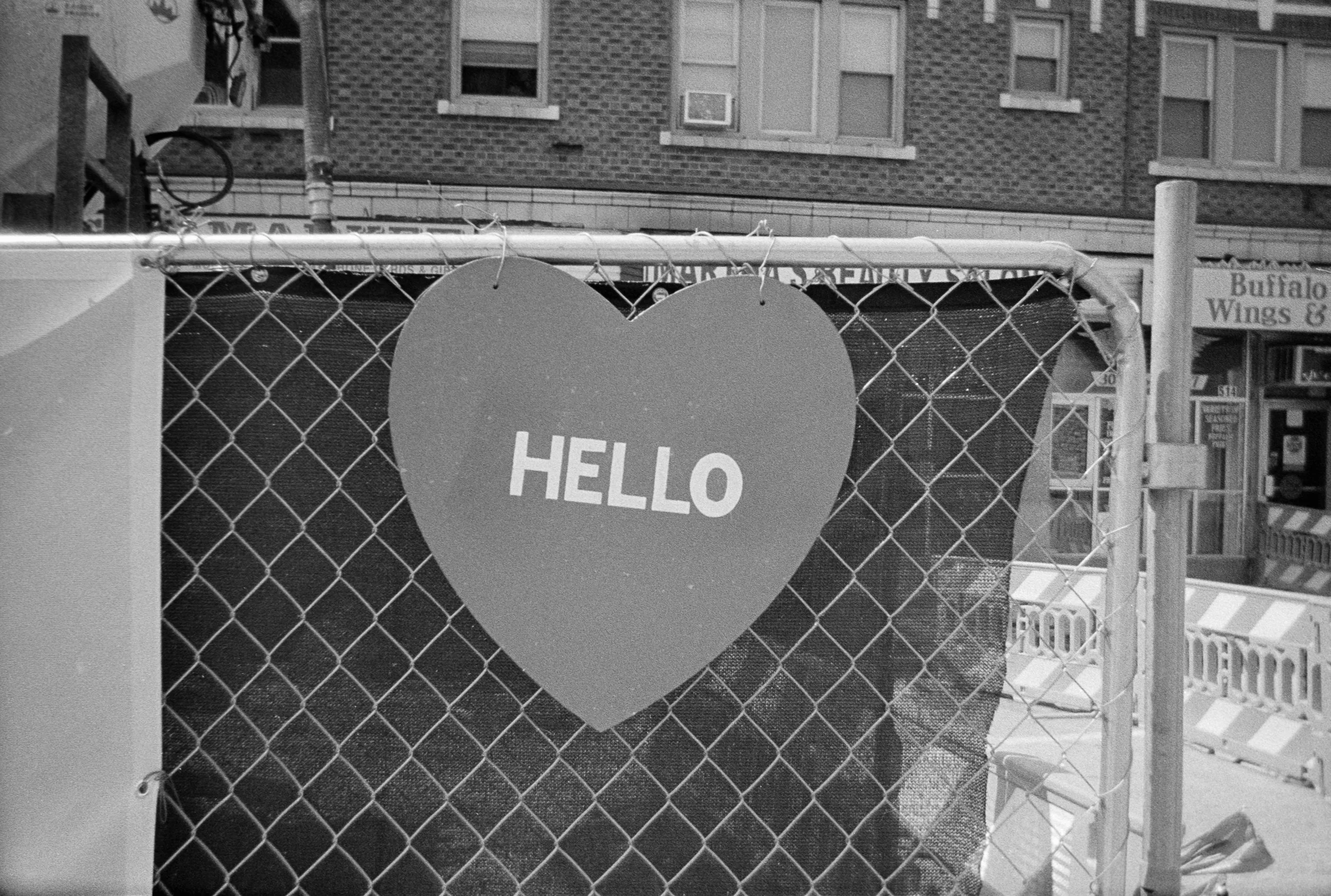 A chain-link fence with a heart-shaped sign that says 'HELLO' in capital letters. In the background, there are buildings and a small storefront sign that reads 'Buffalo Wings &...'