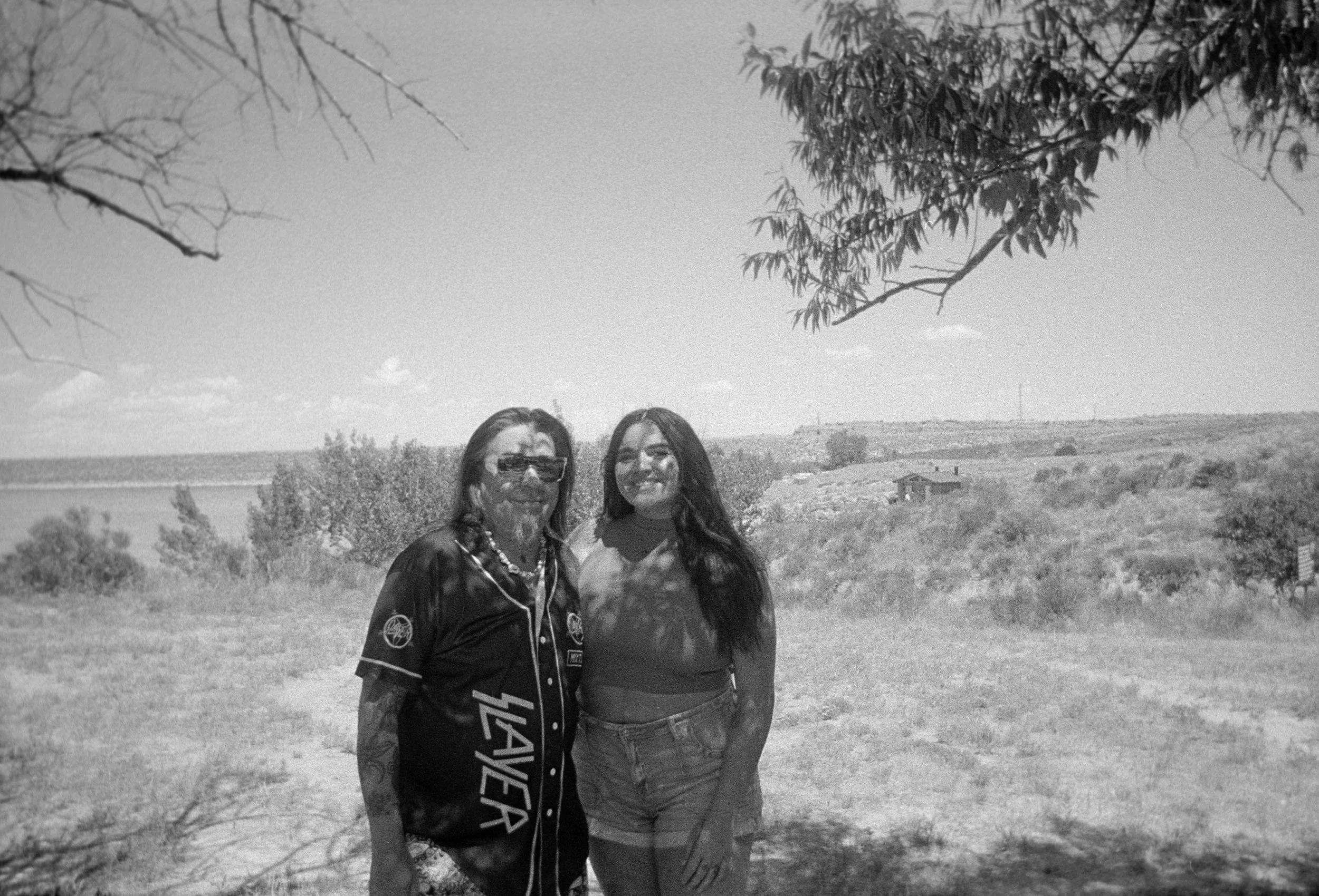 Father and daughter standing outdoors under a tree, smiling at the camera with fields and a few buildings in the background.