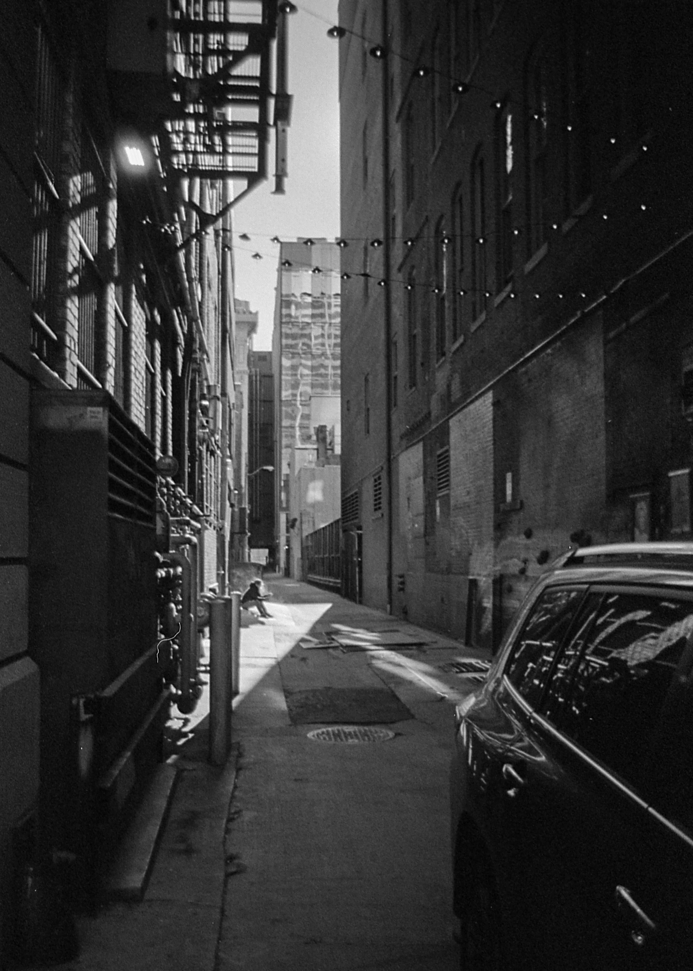 A black and white photo of an alleyway in an urban area, with a car parked to the right, string lights hanging above, a person sitting on the sidewalk in the distance, and tall buildings in the background.