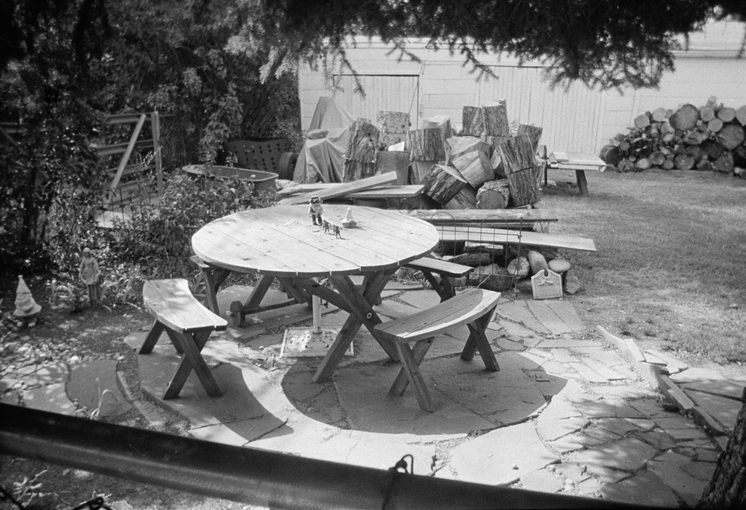 A backyard patio with a wooden picnic table and bench set on a stone circular area. In the background, there is a pile of chopped wood, some stacked logs, and various outdoor garden decorations. A tree branches over the scene, casting shadows.