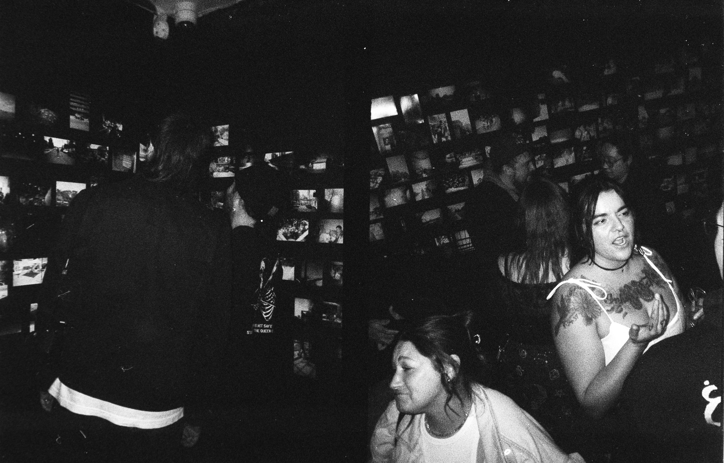 Black and white photo of people socializing in a dimly lit bar or club, with walls decorated with shelves of small framed pictures.  Opening show for Collective.Objective