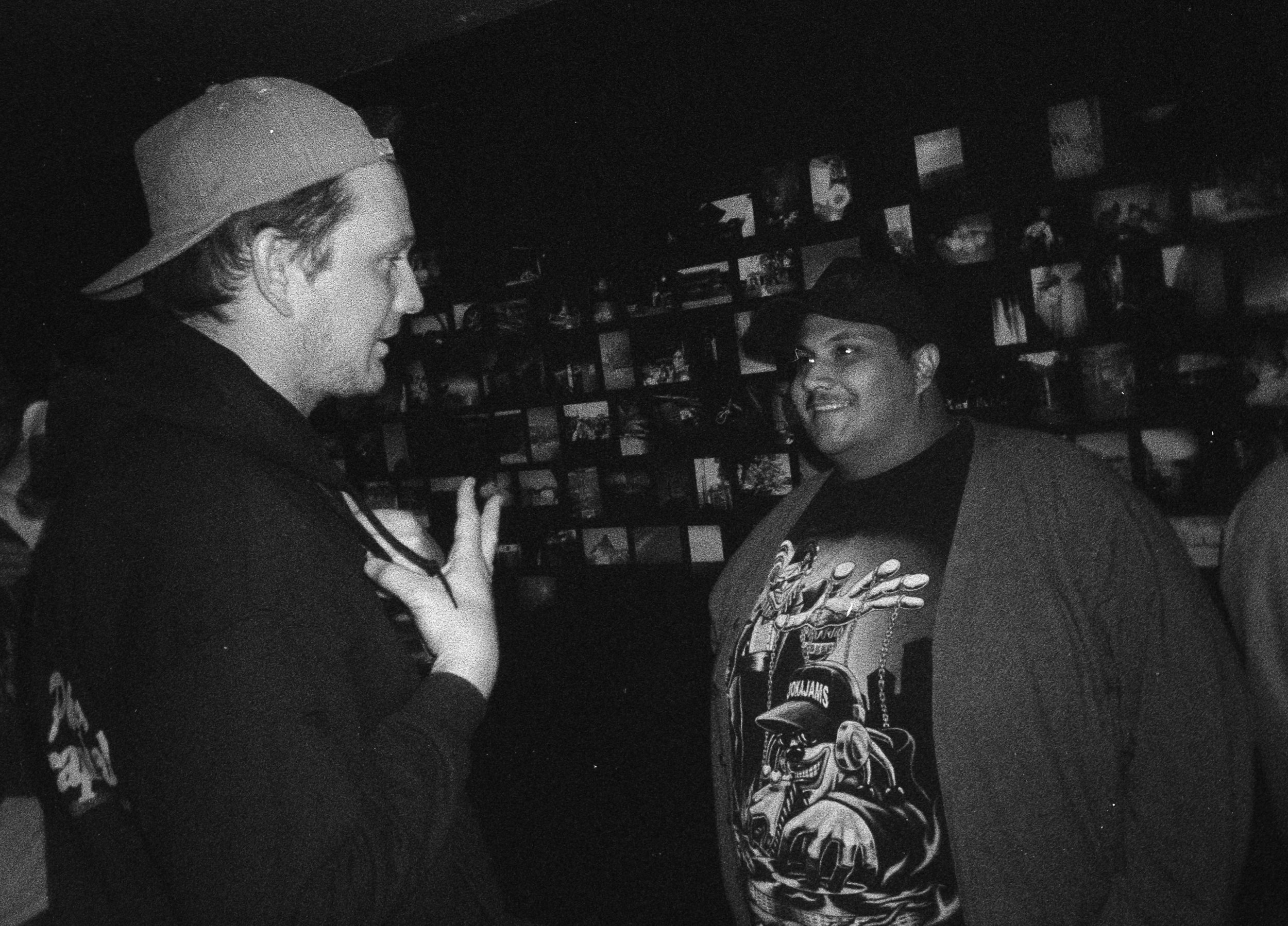 Two men engaged in conversation in a dimly lit room with photo displays on the wall behind them.  Opening for Collective.Objective