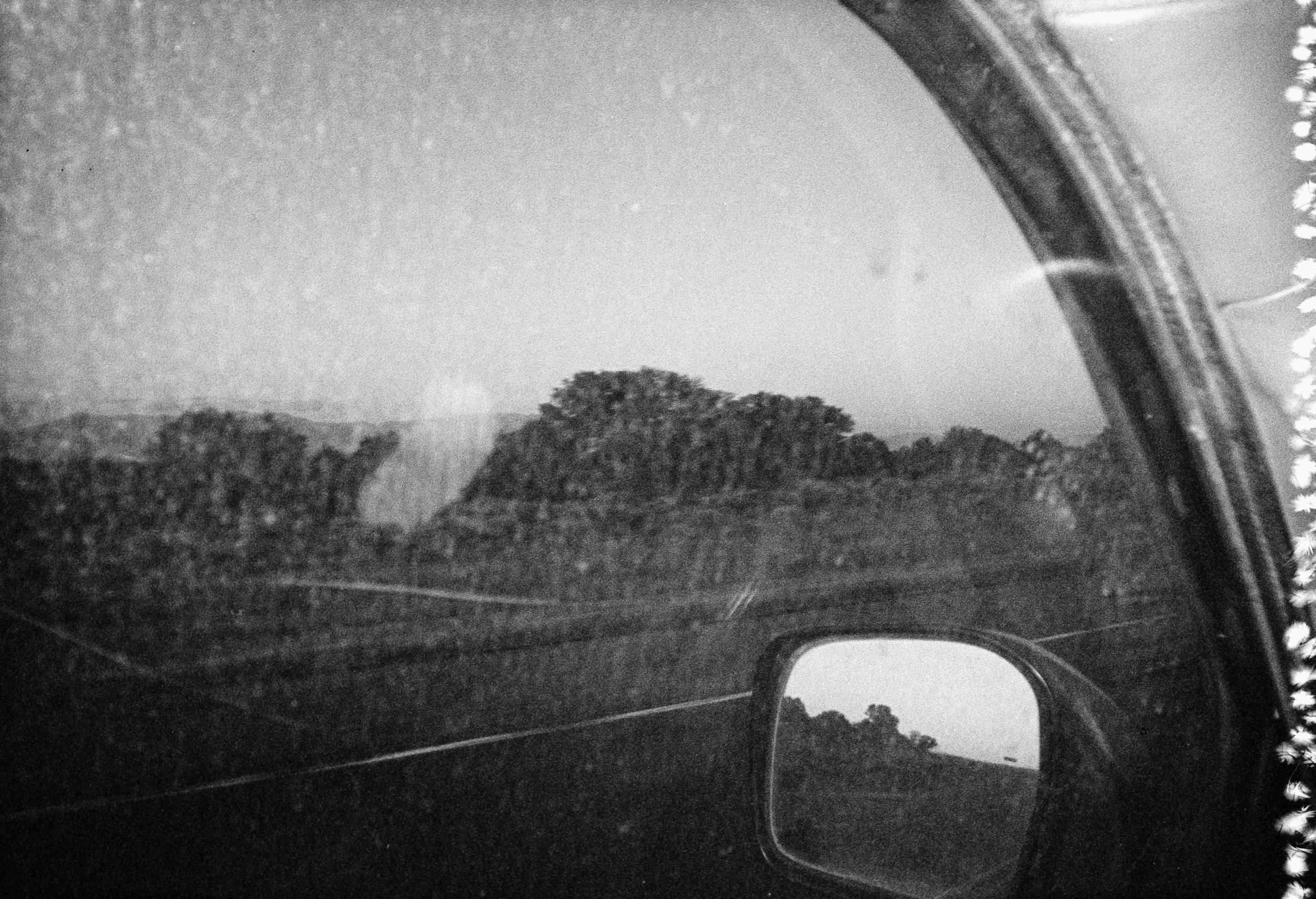 Black and white photograph capturing a landscape viewed through the side mirror of a vehicle. Focus on distant hills or mountains and some trees, with a textured surface of the mirror showing signs of wear or dust.