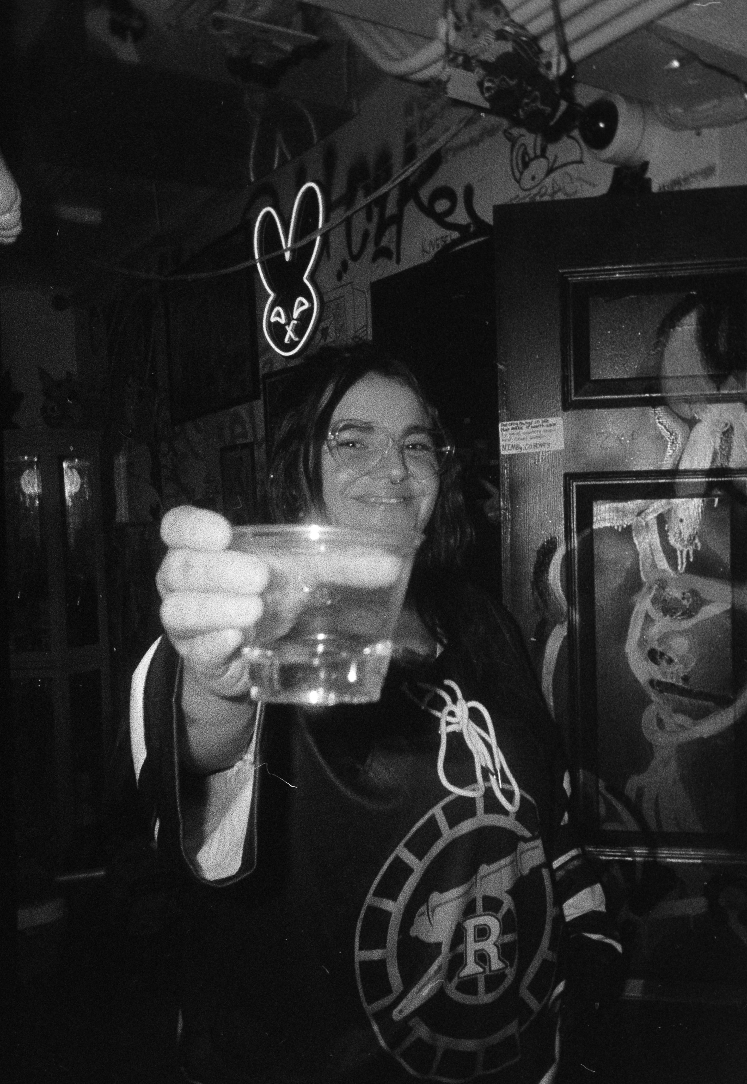 A woman wearing glasses and a hockey jersey is smiling and holding up a glass of clear drink towards the camera in a dimly lit bar or club with graffiti and art on the walls, including a neon bunny sign in the background. Opening for Collective.Objec