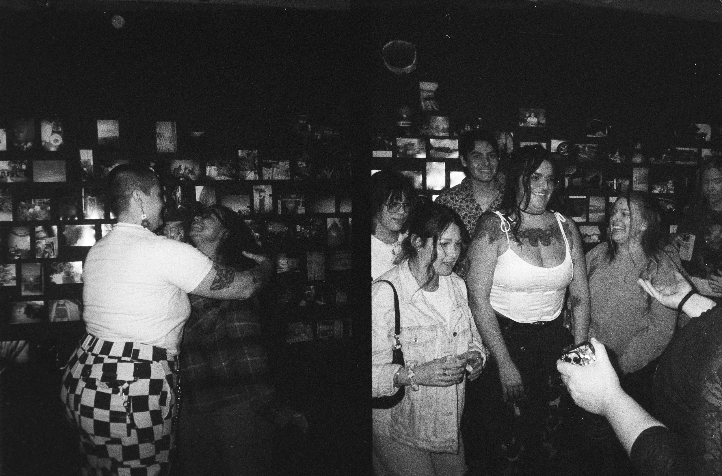 A group of people gathered in a dimly lit space with a wall full of photos in the background, some smiling and interacting.