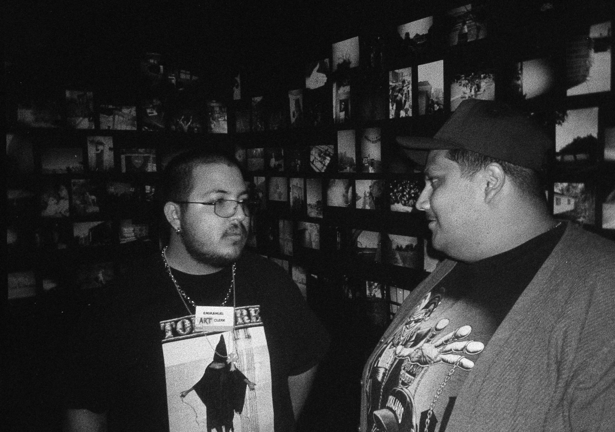 Two people talking in front of a wall display of framed photos, one wearing glasses, a name tag, and a picture T-shirt, the other wearing a hat and a T-shirt with a graphic design.  Opening for Collective.Objective