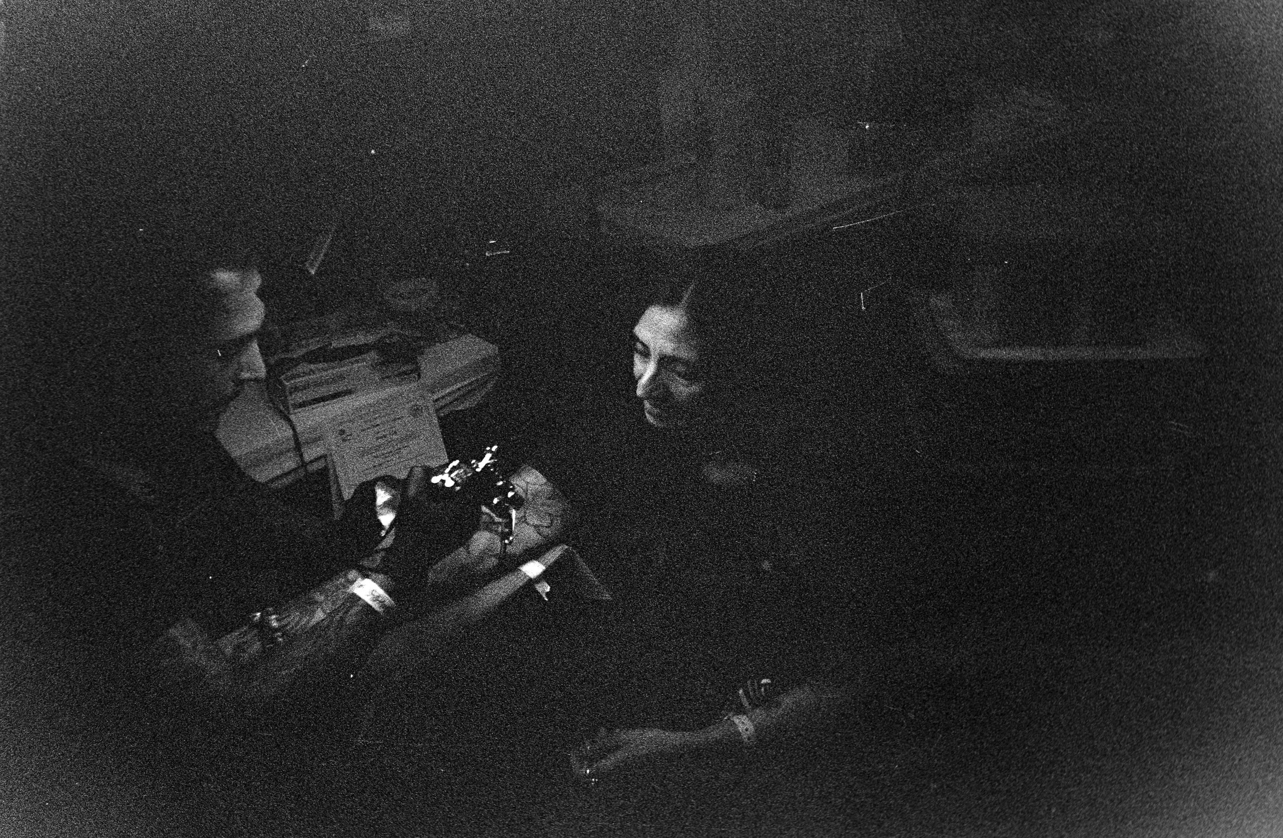 Two people sitting in a dark room, one is tattooing the other.