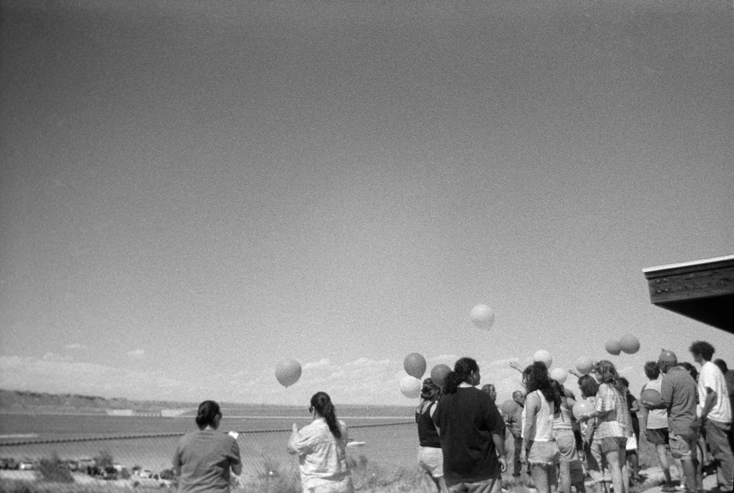 A group of people gathered outdoors by a body of water, with balloons floating in the sky during daytime.