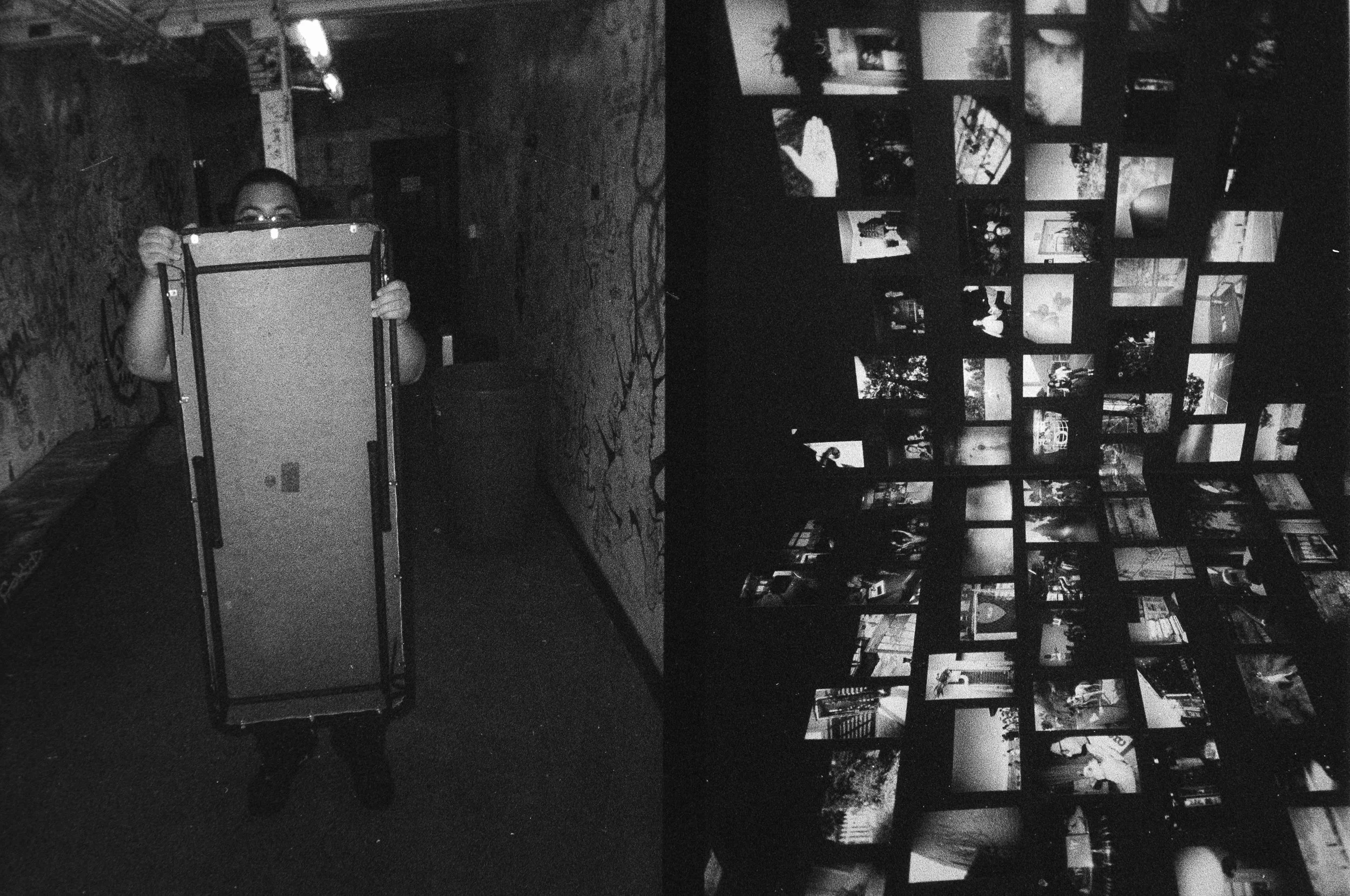 A person holding an empty tray in a dimly lit room, with a collage of photographs displayed on the wall to the right. Opening show for Collective.Objective