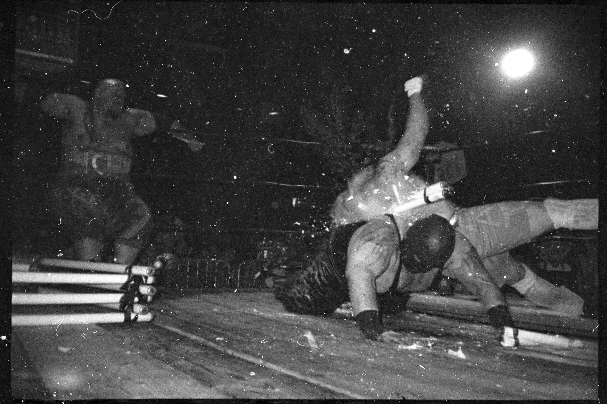 A black and white photograph of a wrestling match with a fighter falling to the mat, while another fighter and a referee look on. The scene is intense, with the wrestler on the ground attempting to recover and the referee reaching out. 