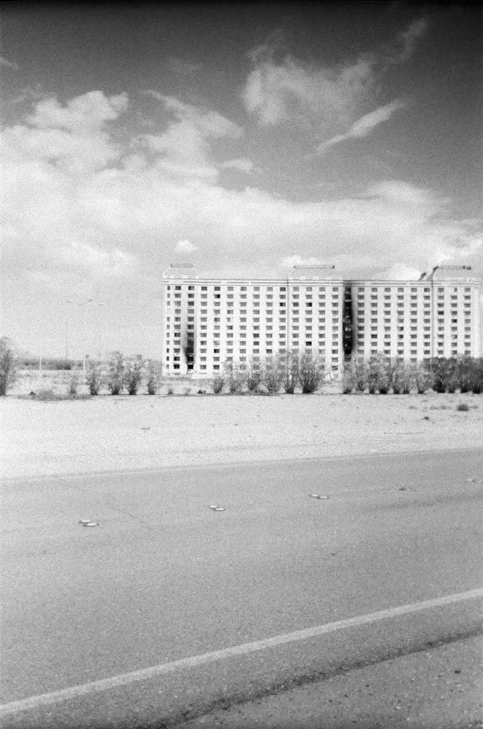 Black and white photo of a large, burned out hotel building with many windows, trees in front, and sky with clouds above.