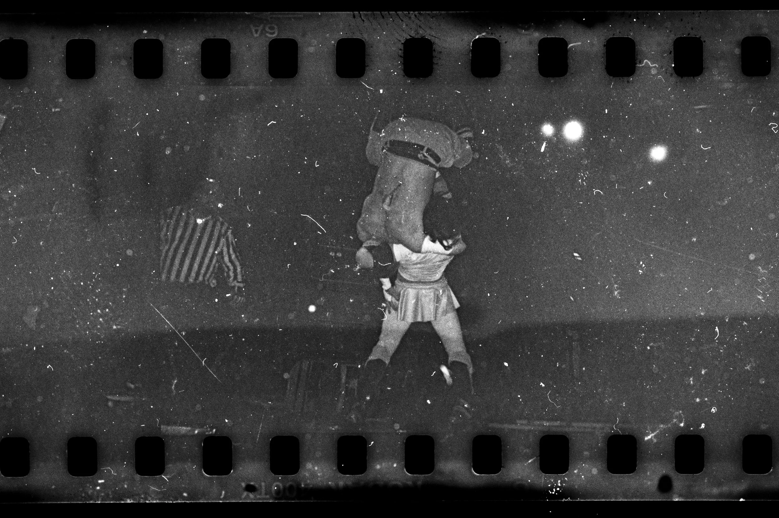 Black and white photograph of a wrestler carrying another person on their shoulders, with a referee nearby. The scene appears lively with bright lights and a grainy texture. film developed in pig's blood