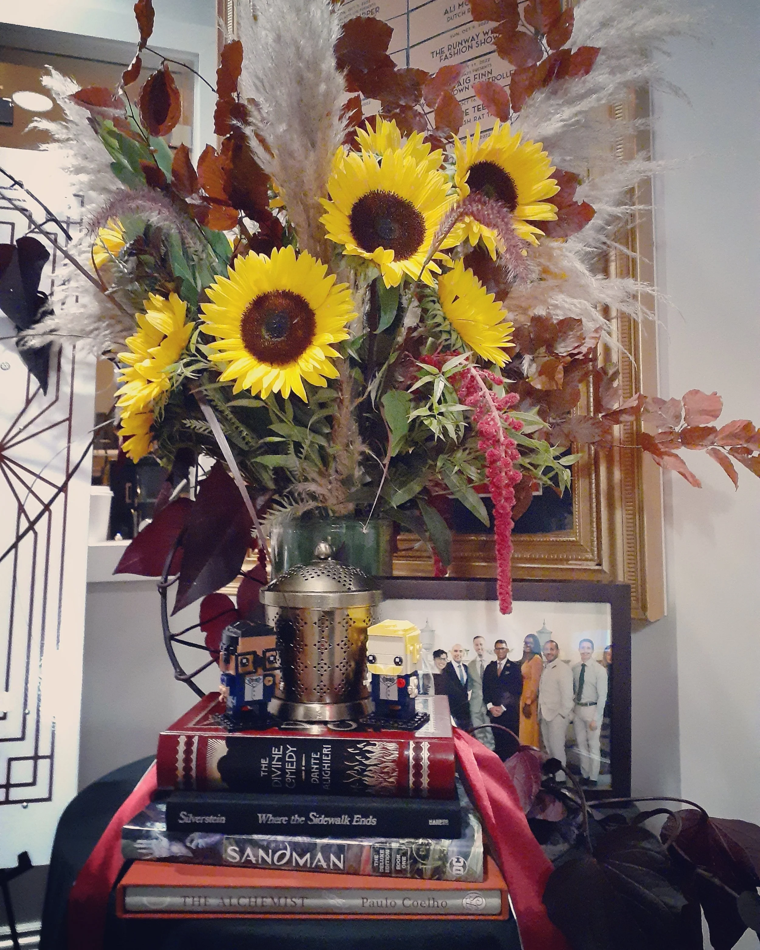 A large bouquet of sunflowers, autumn leaves, and dried grasses in a vase, placed on a table with books and small decorative figures, with a framed photograph and wall decorations in the background.
