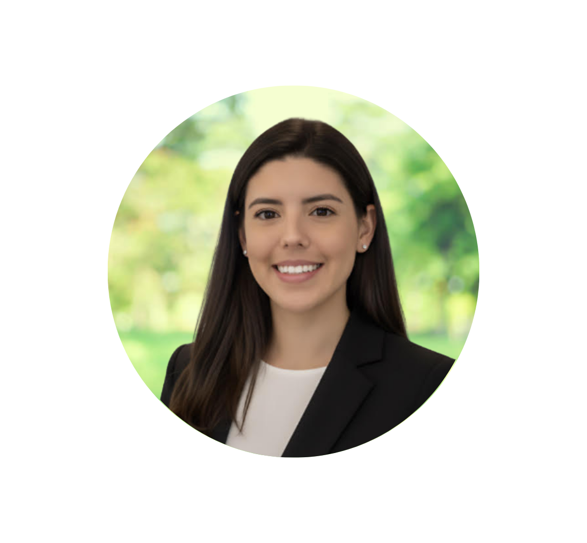 Portrait of a young woman with long dark hair wearing a black blazer and white top, smiling, with a blurred green outdoor background.