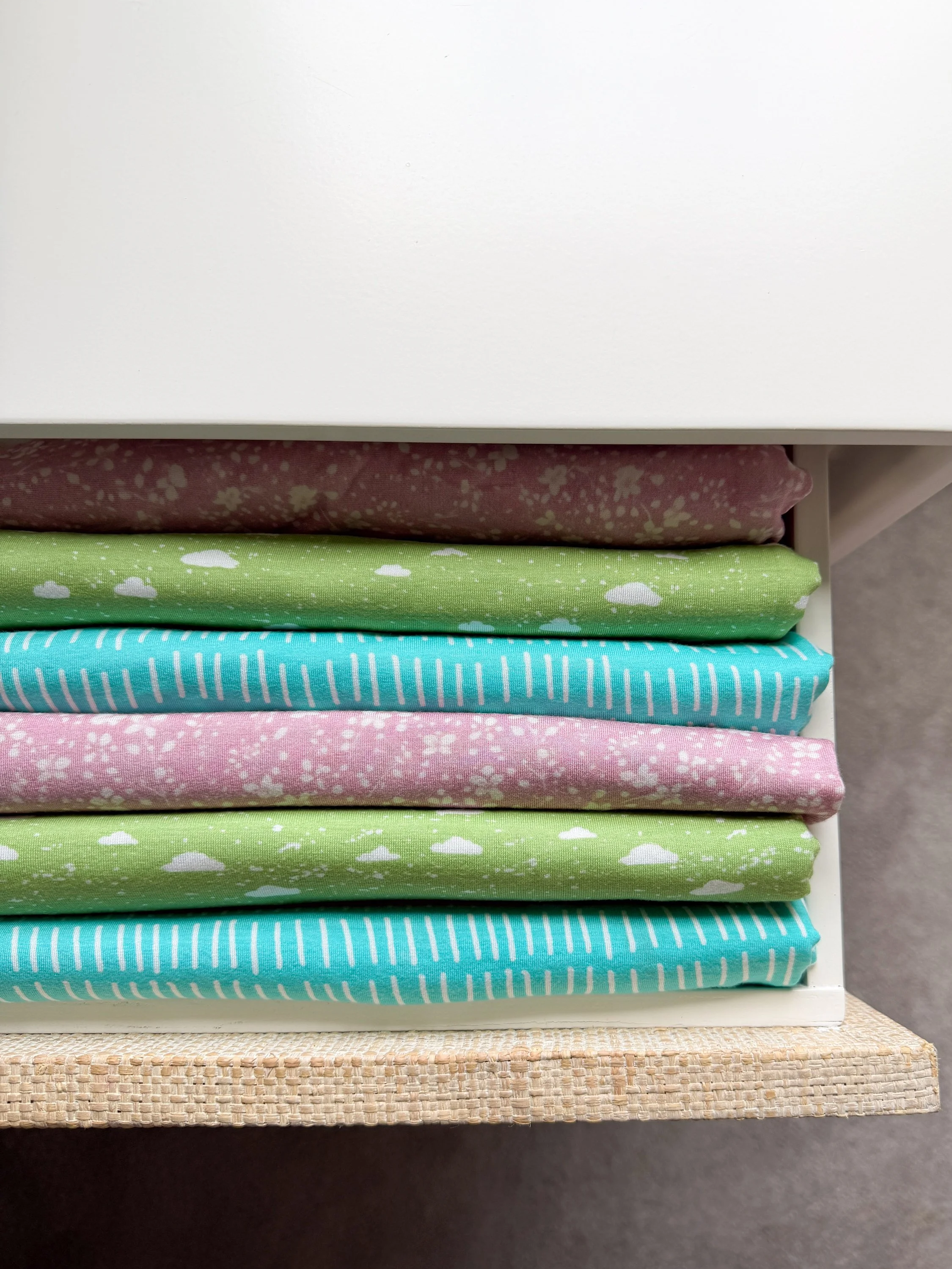 Stacked folded fabrics in pink, green, light blue, and pink with white patterns sitting on a white shelf with a woven rattan surface below.