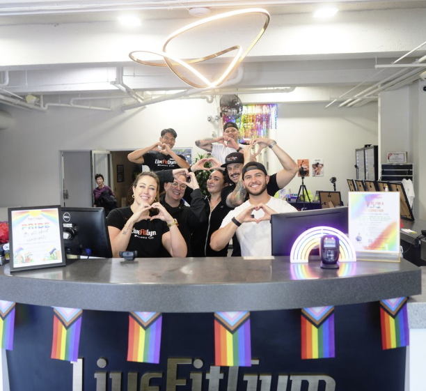 Group of seven people at a registration desk making heart signs with their hands, decorated with rainbow Pride flags, in a modern, colorful event space.