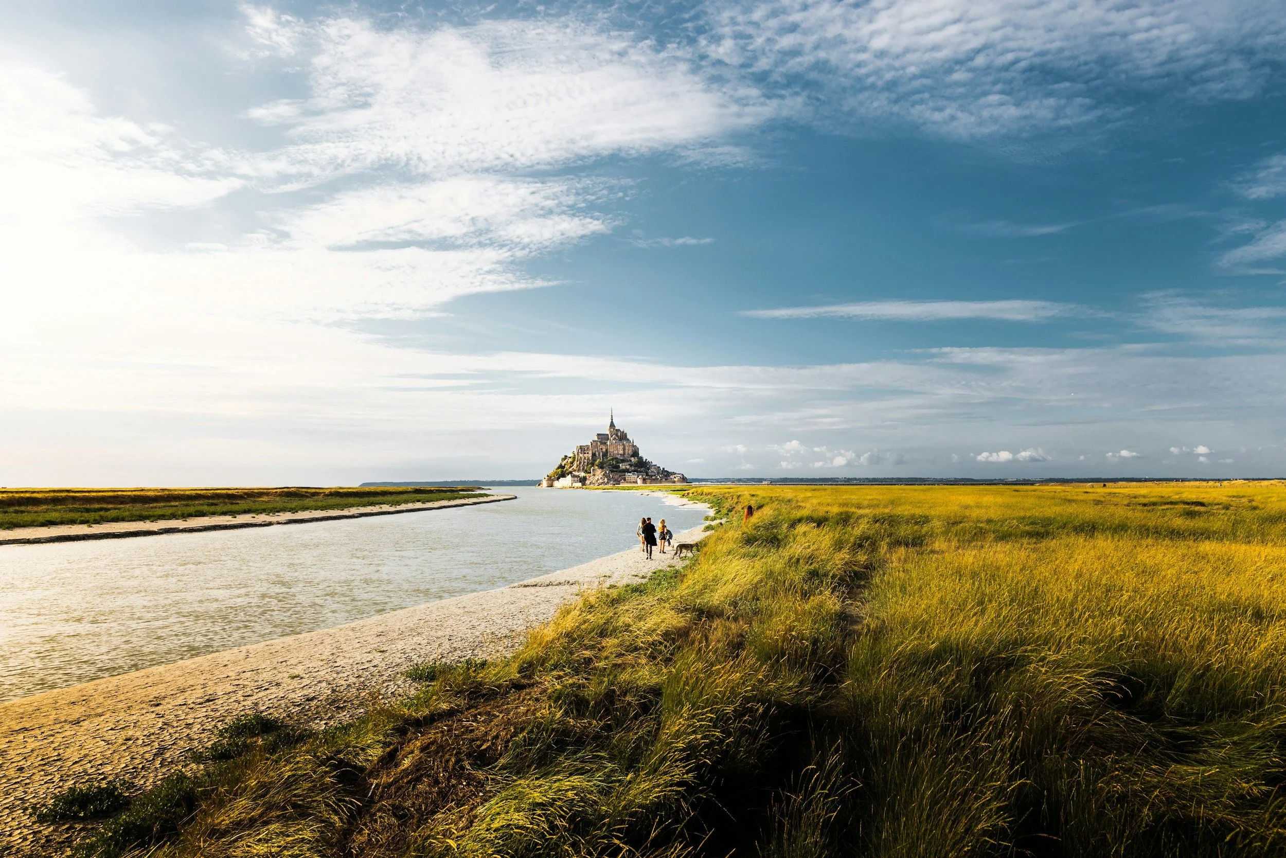 eite Landschaft mit einem Flusslauf und gelbem Gras im Vordergrund, im Hintergrund thront die Klosterinsel Mont-Saint-Michel unter einem blau-weißen Wolkenhimmel.