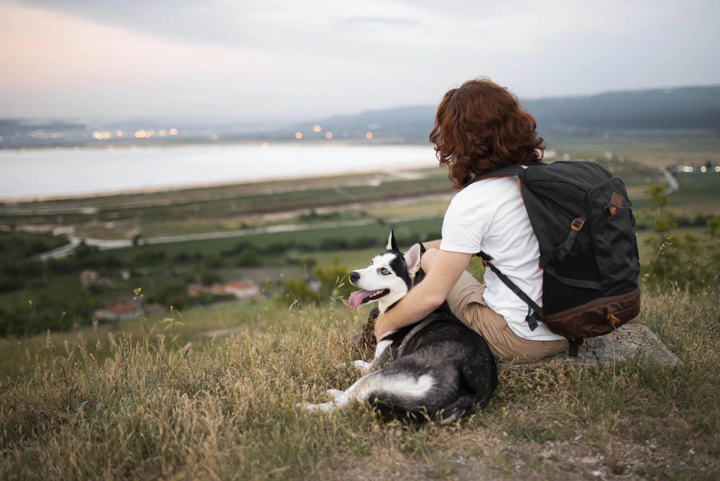 Person mit Rucksack sitzt mit einem Husky auf einem Hügel und blickt auf einen See und die umliegende Landschaft.