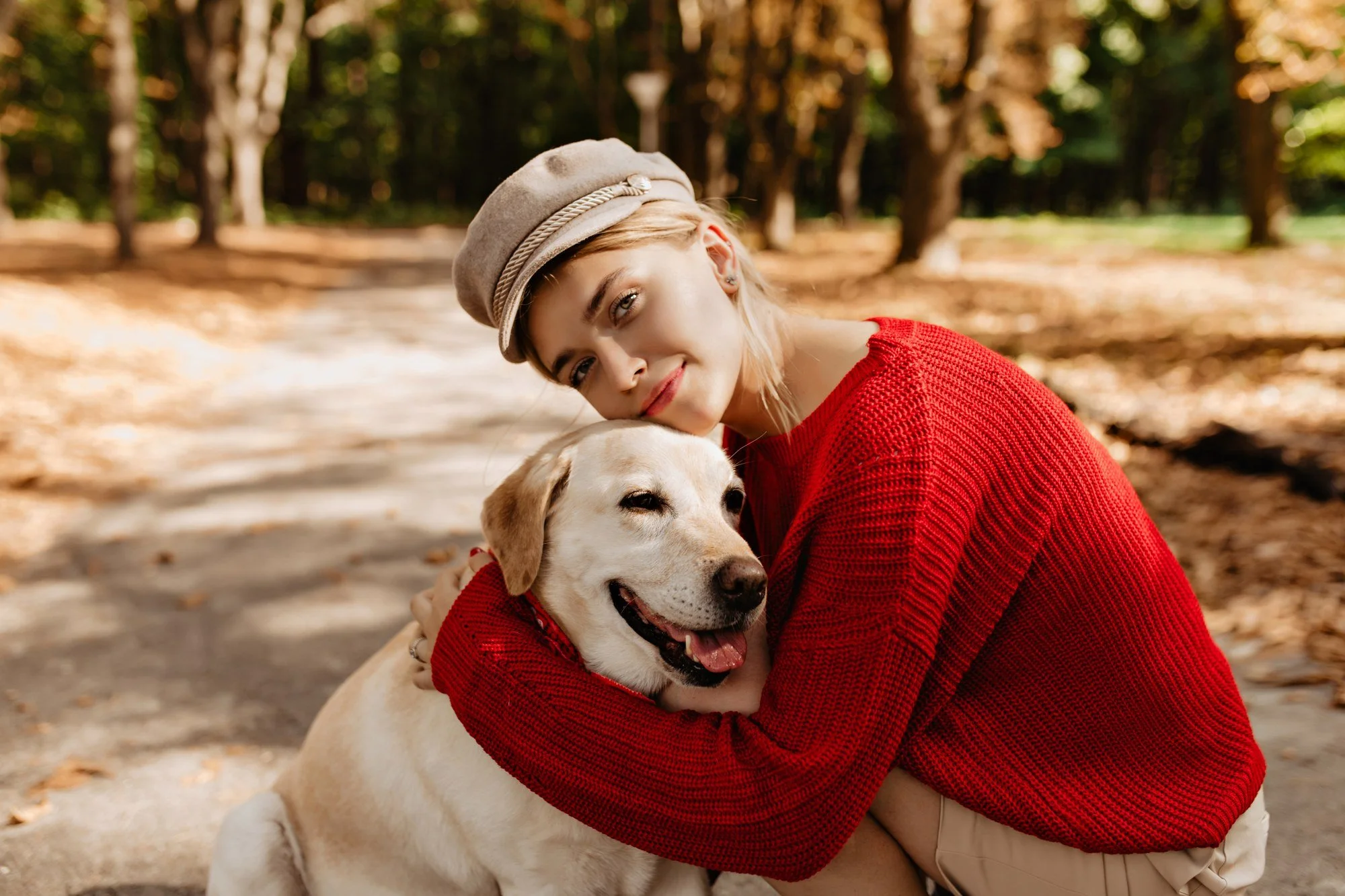Frau mit heller Mütze und rotem Pullover umarmt einen Labrador im herbstlichen Park.