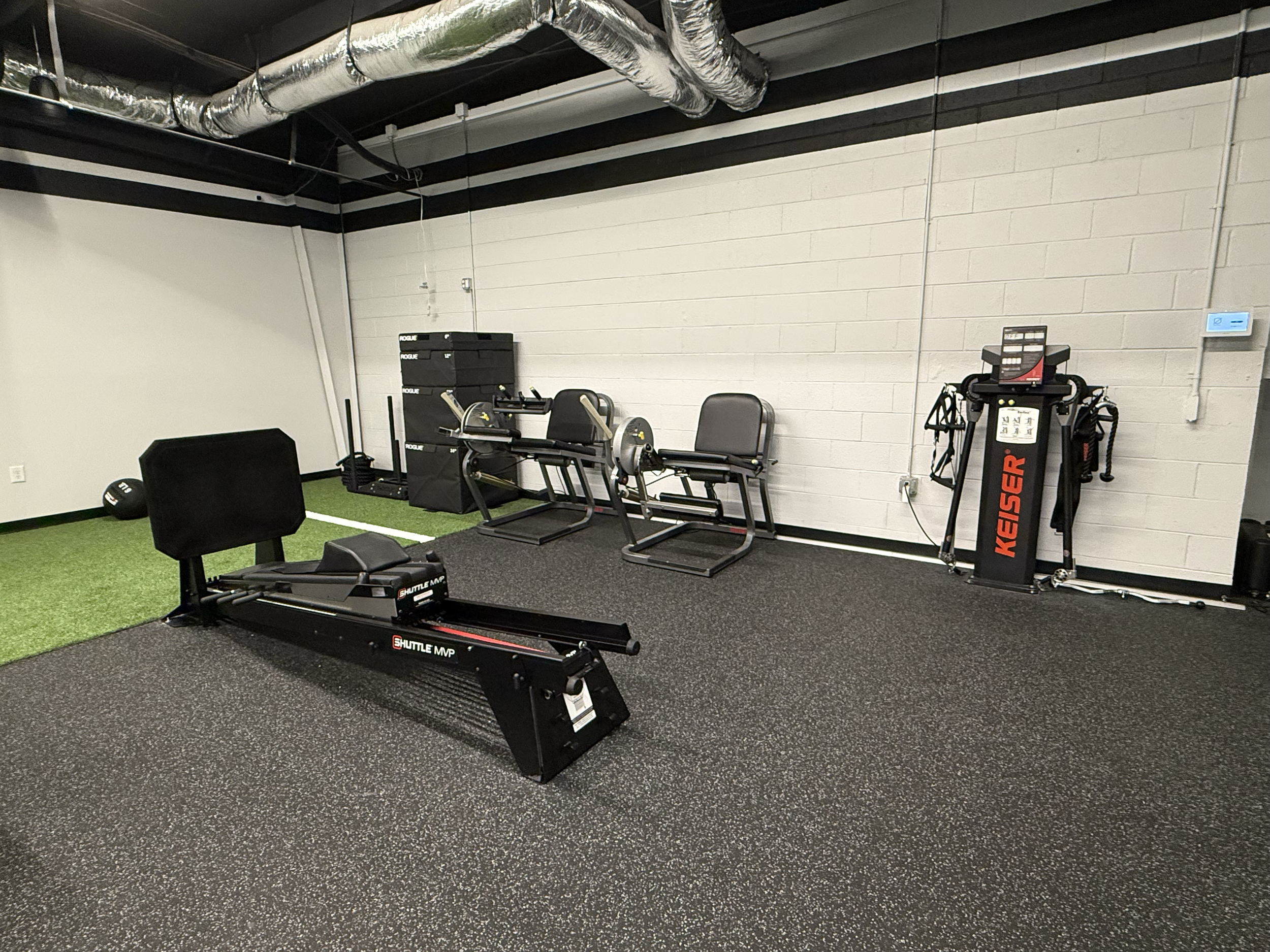 A gym with black and gray equipment on a rubber flooring, including a rowing machine, a weightlifting machine, a chair, a power rack, and other fitness equipment. The gym has a white brick wall and a ceiling with exposed ductwork.