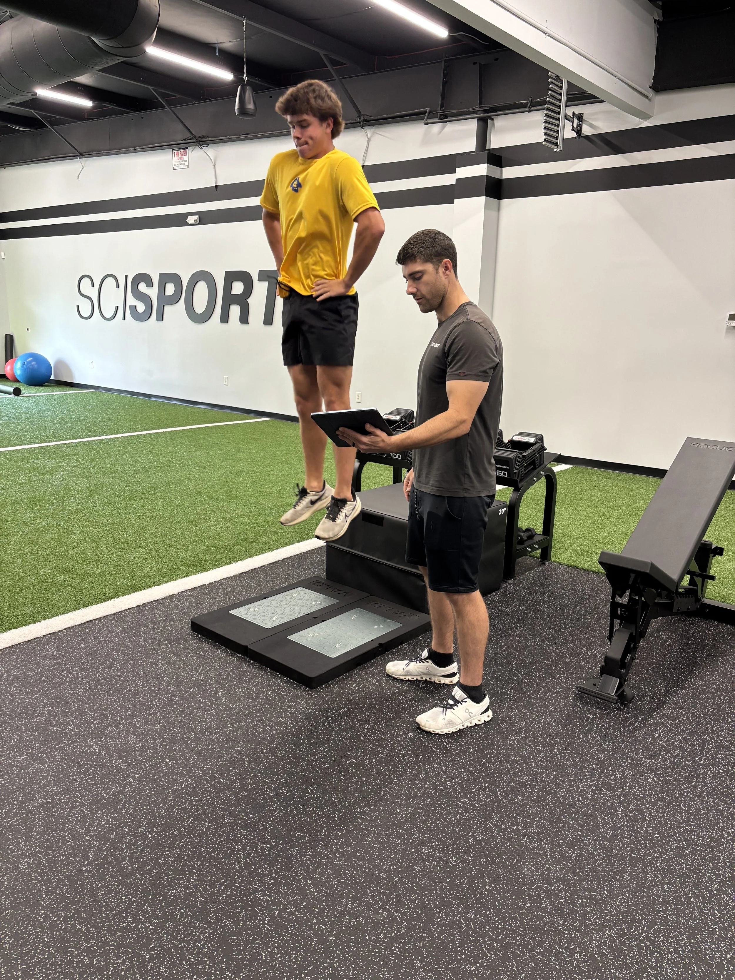 A man is instructing a young man performing a jump during a fitness assessment at the SciSport gym, with the young man mid-air and the instructor holding a tablet.