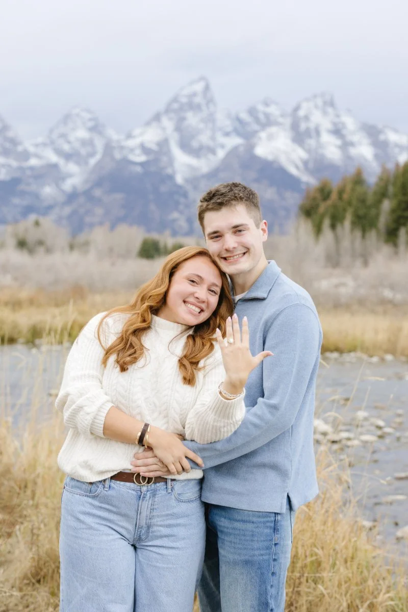 Smiling couple showing off an engagement ring after their Grand Teton proposal with the Teton mountain range in the background