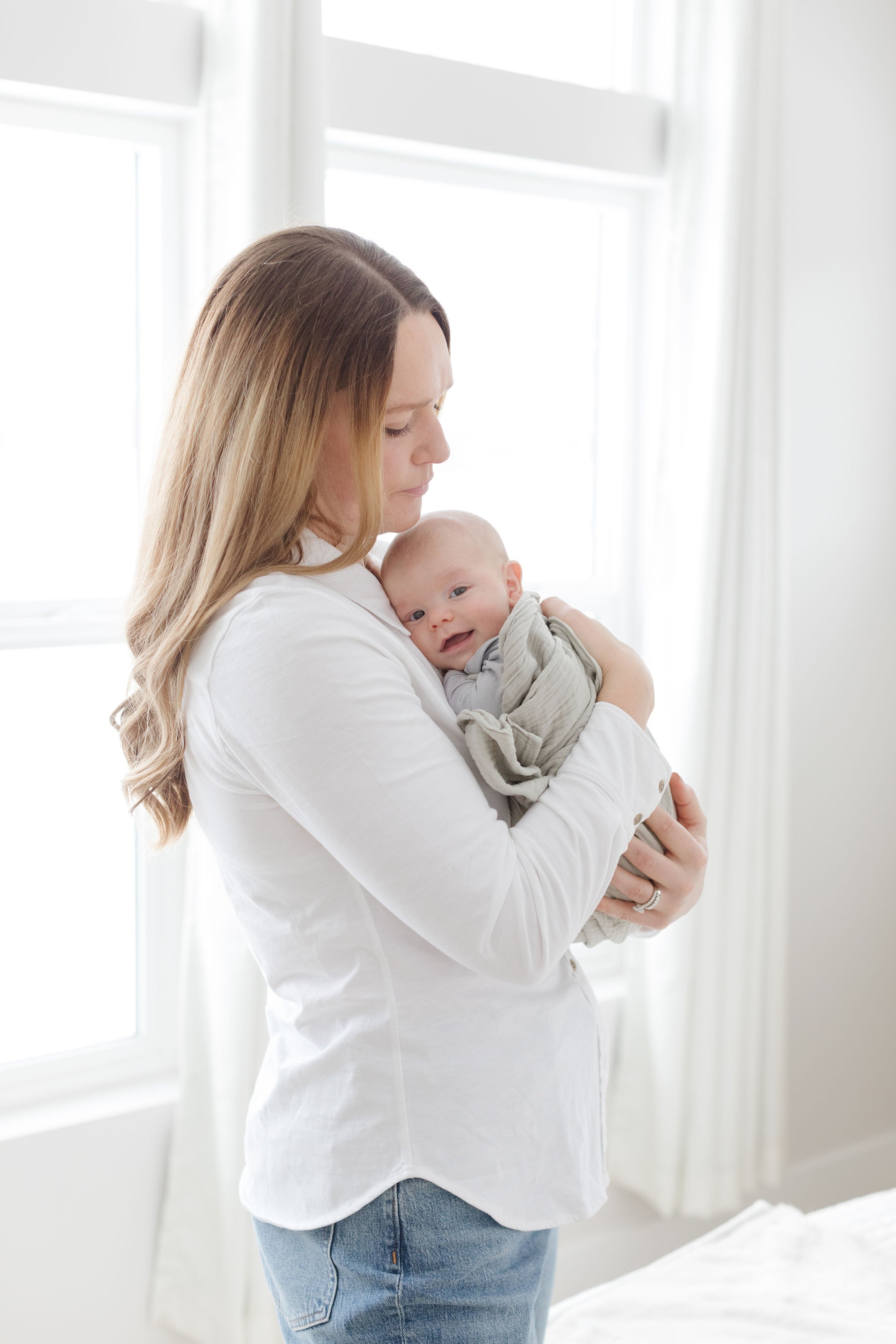  Mom holds baby as a newborn photographer in Jackson Hole captures their photograph in front of a bright window. 