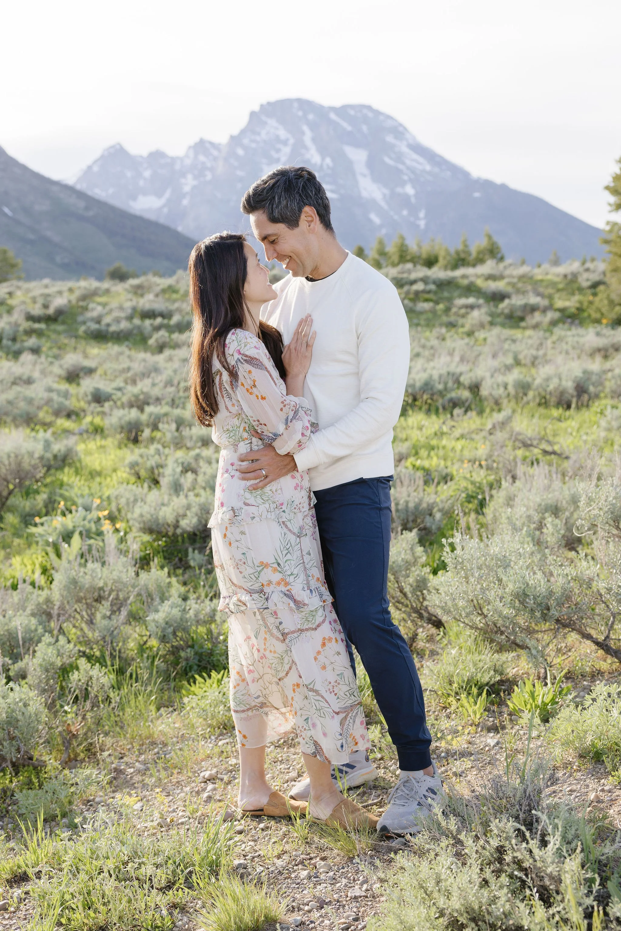  Couple pose for photo in the Grand Tetons, surrounded by greenery and mountain views. 
