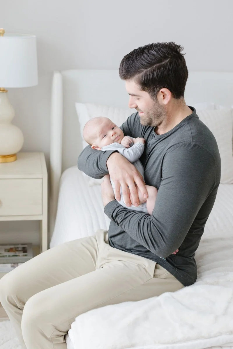  Dad holds newborn baby as he sits in bedroom. 