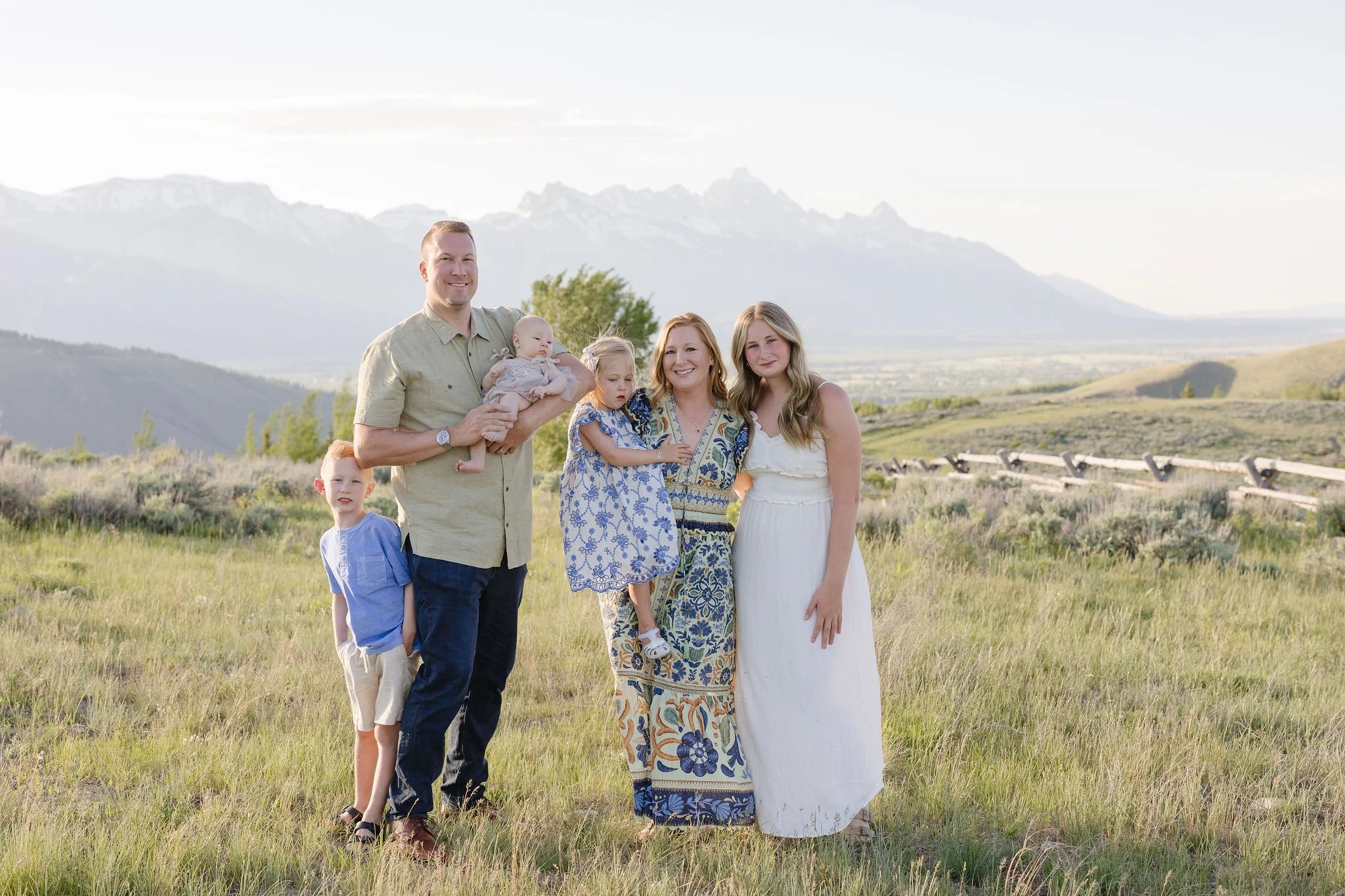 Family stands together in a grassy field with the Grand Teton sunrise coming up behind them.