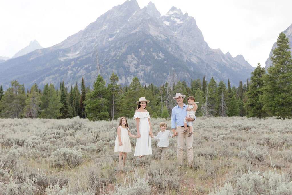  Family stands together holding hands in the Tetons as a photographer captures their family photos. 