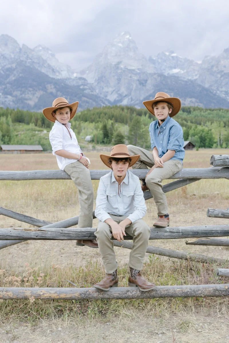  Three brothers in cowboy hats sitting on a wooden fence with the Grand Tetons in the background 