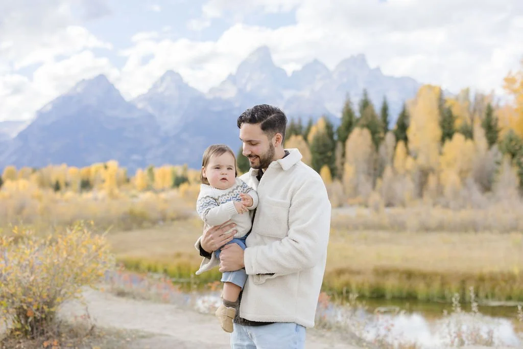 Father holds baby with Grand Tetons in the backyard - noting fall is the best time to visit Grand Teton National Park.